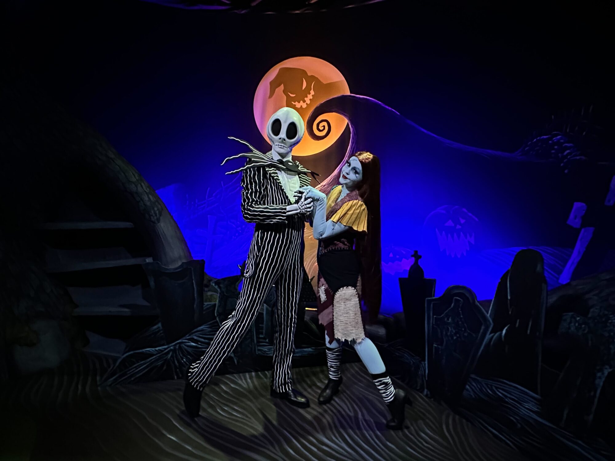Two costumed characters stand in a Halloween-themed setting with a not-so-scary, spooky moonlit background and cheerful pumpkin faces.