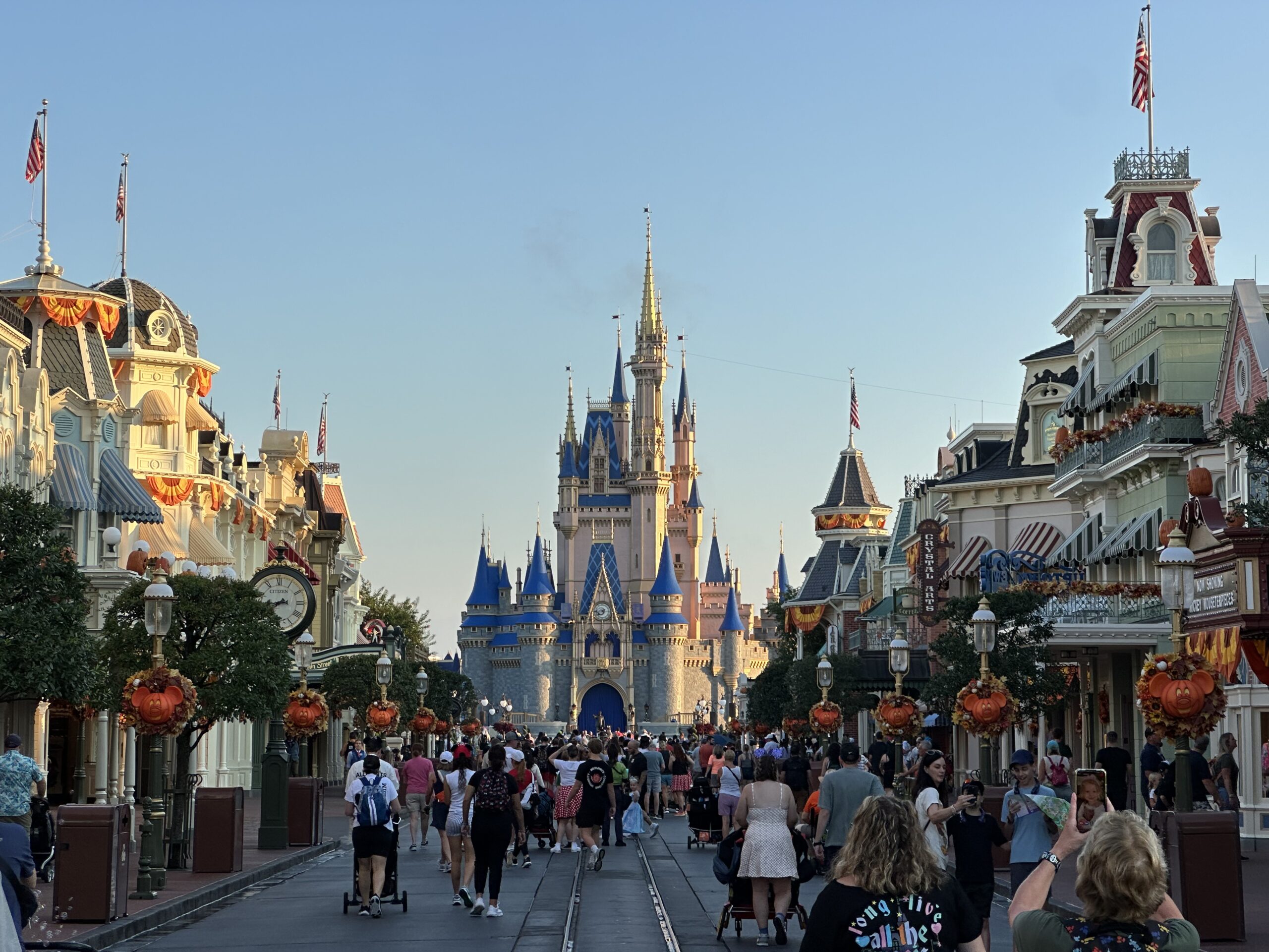 A crowd of people walk down Main Street towards Cinderella Castle at Magic Kingdom in Disney World. The street is decorated with pumpkins and banners.