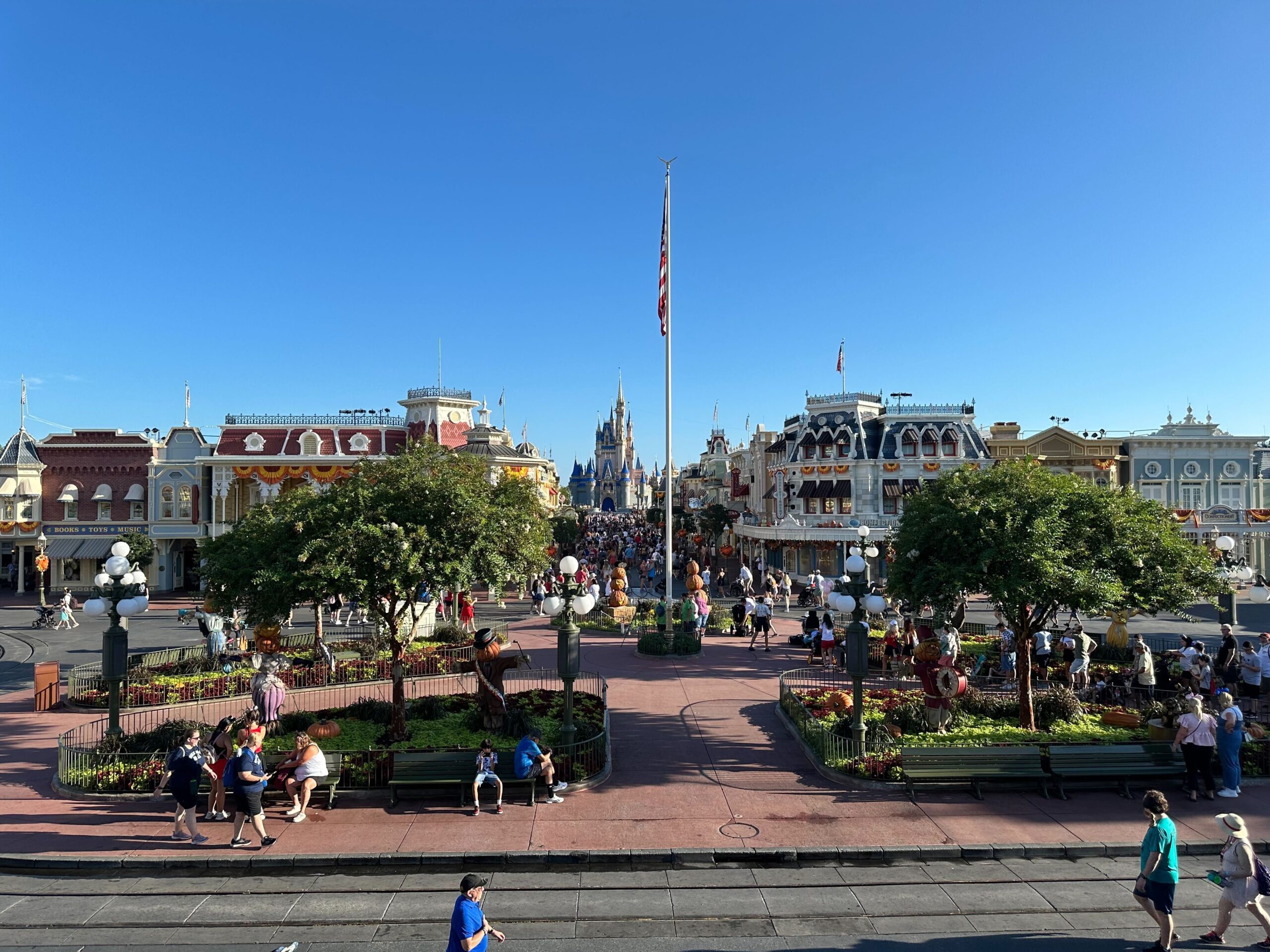Wide-angle view of Magic Kingdom's Main Street, featuring numerous visitors, themed buildings, and a distant castle under a clear blue sky.