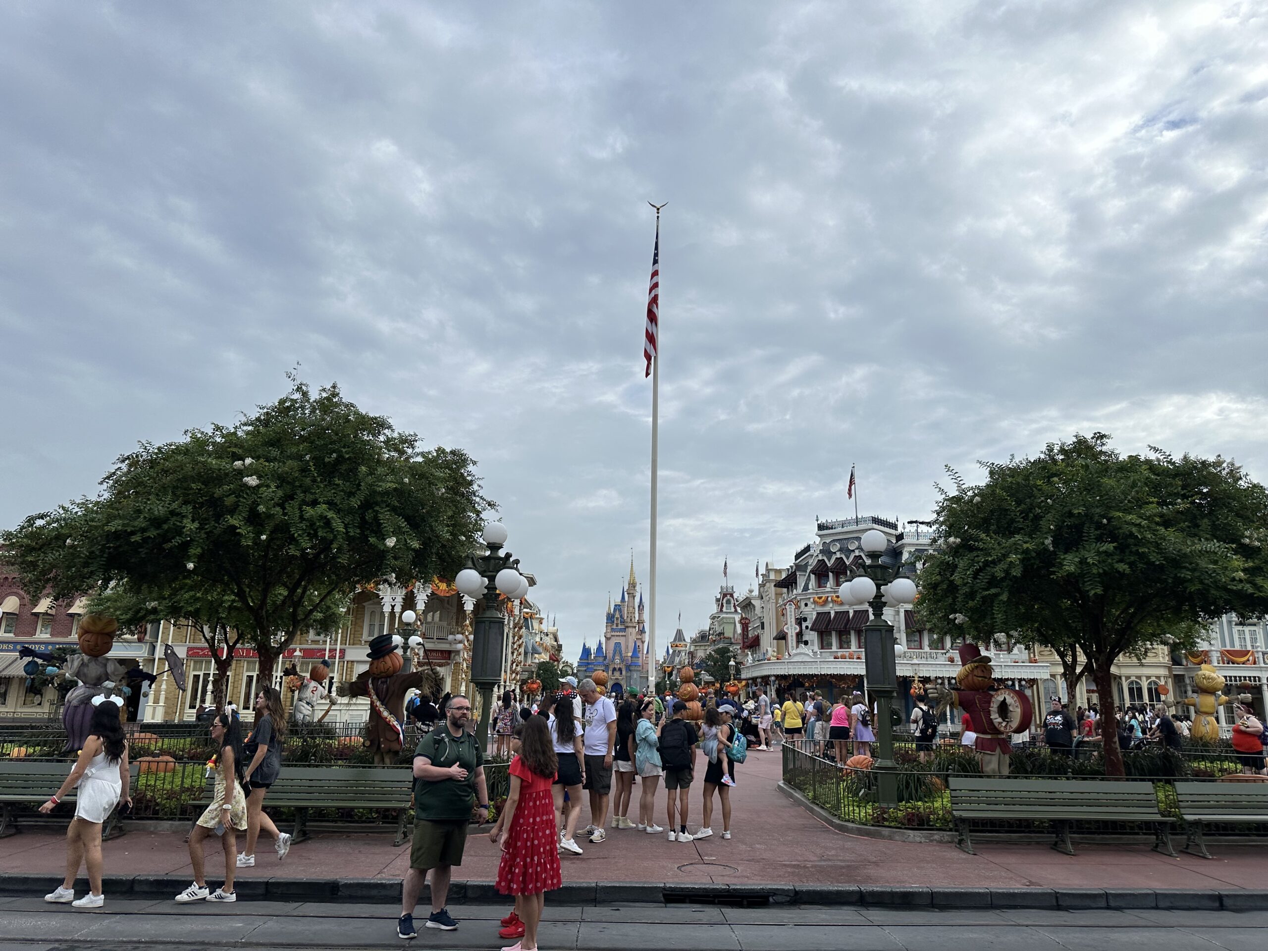 People are gathered in an amusement park area with a flagpole in the center and a castle in the background under a cloudy sky. Magic Kingdom, Cinderella Castle in the center behind the flag pole.