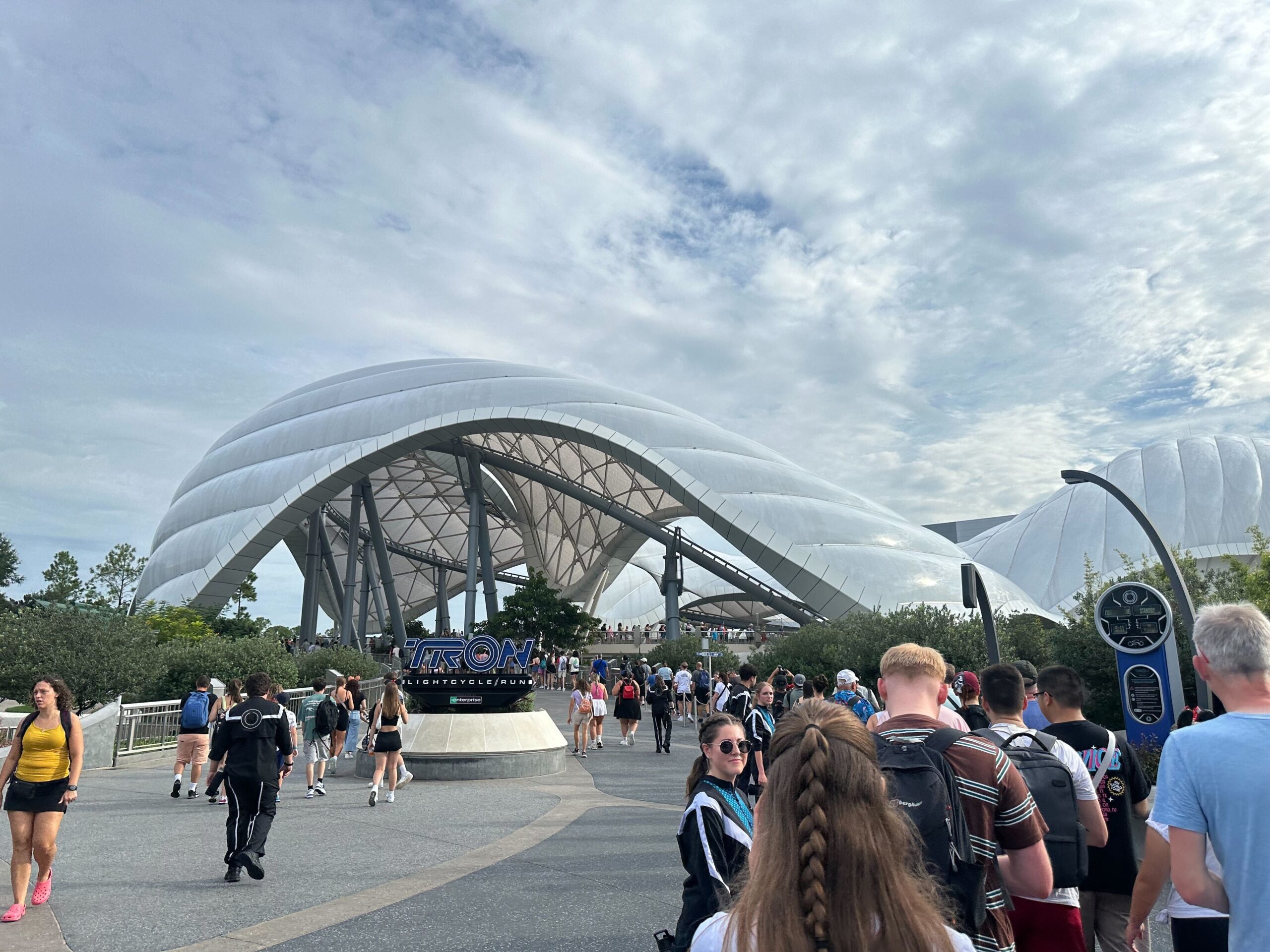 People walking towards a large, futuristic structure with white, curved roof and "TRON" signage in front. The sky is partly cloudy.