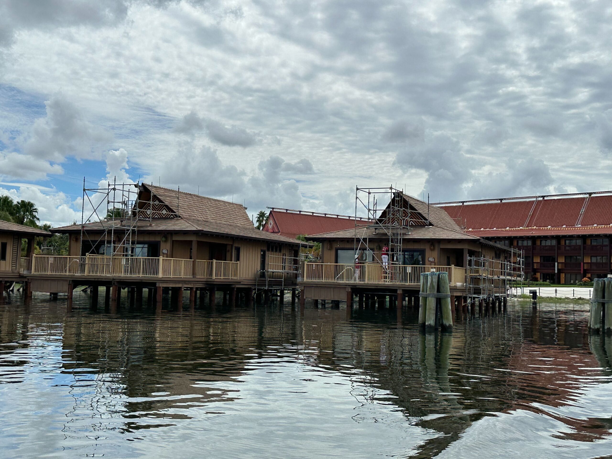 Overwater bungalows on stilts undergoing construction, with scaffolding visible around the buildings, set against a cloudy sky and a larger building in the background.