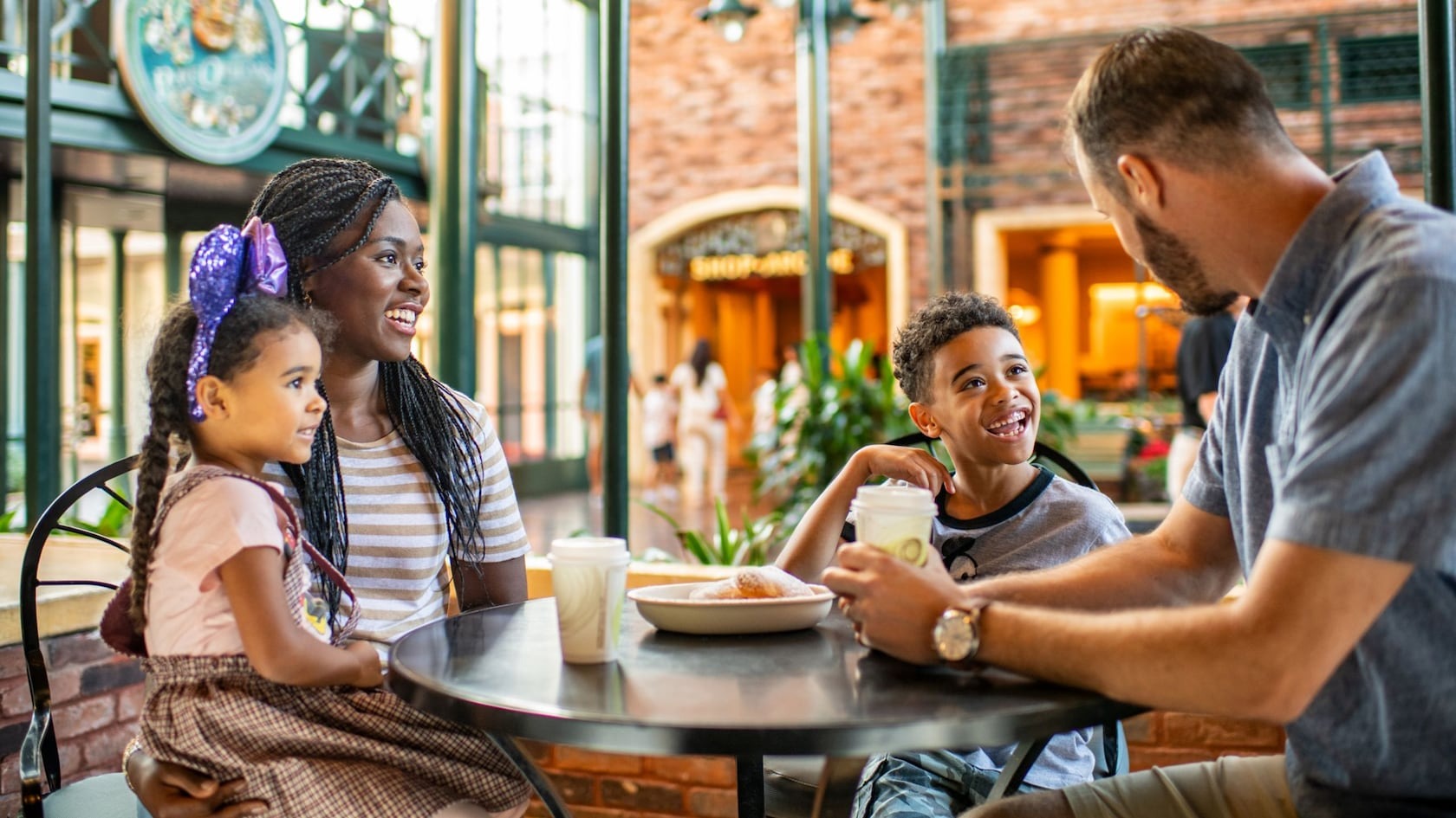 A family of four, consisting of two adults and two children, sits at a café table smiling and talking while having drinks and a pastry. The setting appears to be indoors with a shopping mall atmosphere, reminiscent of the cozy ambiance at Disney's Port Orleans Resort.