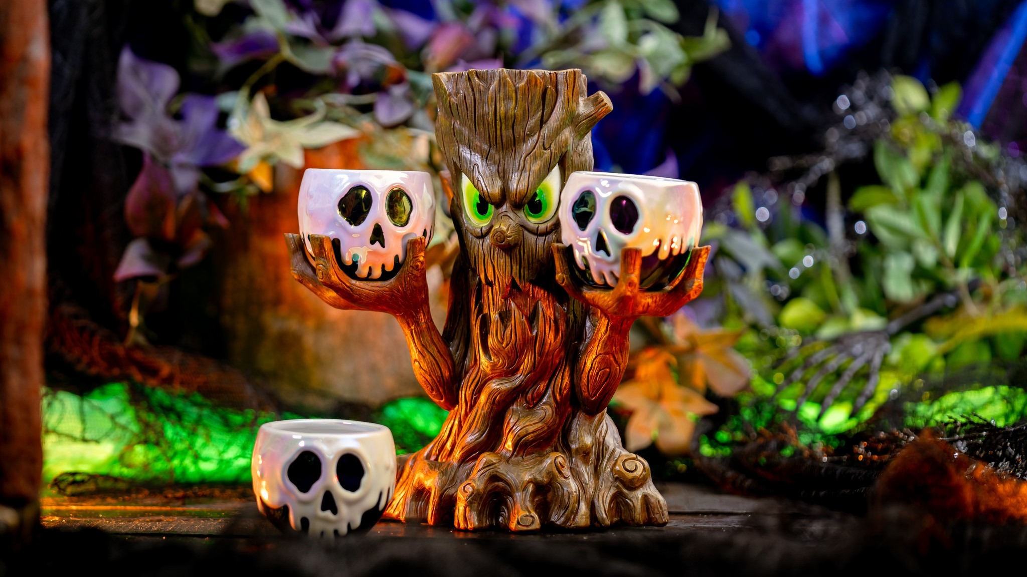 A tree-shaped holder with green eyes and outstretched branches holds three skull-shaped container cups against a backdrop of colorful foliage and moss, much like an auto draft of nature’s own design.