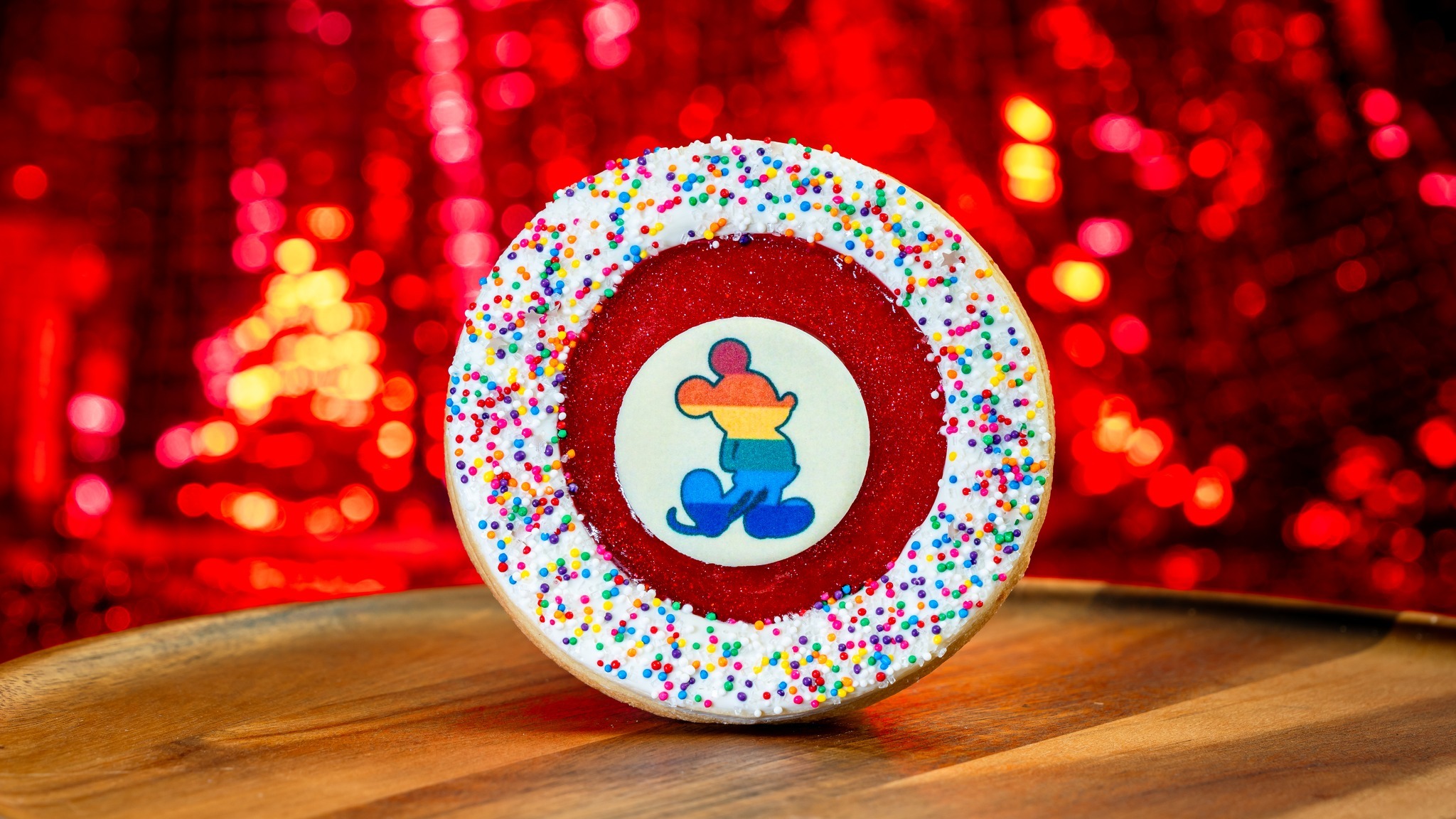 A circular cookie with white icing and colorful sprinkles has a red center featuring an illustration of a character with rainbow colors. The background is a vibrant, glittery red surface.