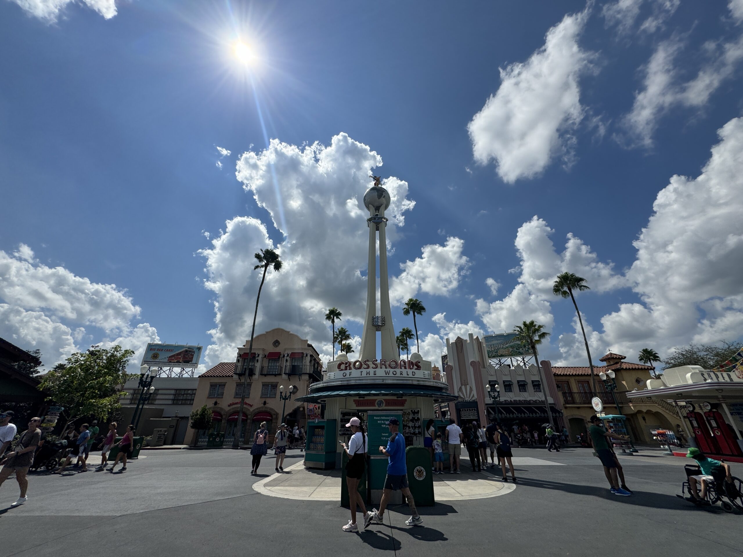 Wide-angle view of Disney's Hollywood Studios entrance under a bright sun with scattered clouds. People are milling around, and palm trees are visible in the background, capturing the vibrant atmosphere in this 2024 photo report.
