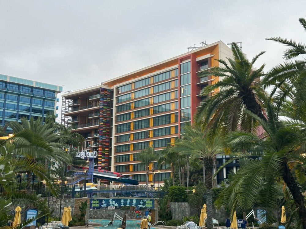 A modern, multi-story hotel with colorful exterior accents and large windows, surrounded by palm trees and facing a pool with yellow umbrellas. In the background, a sign reads "dayland.