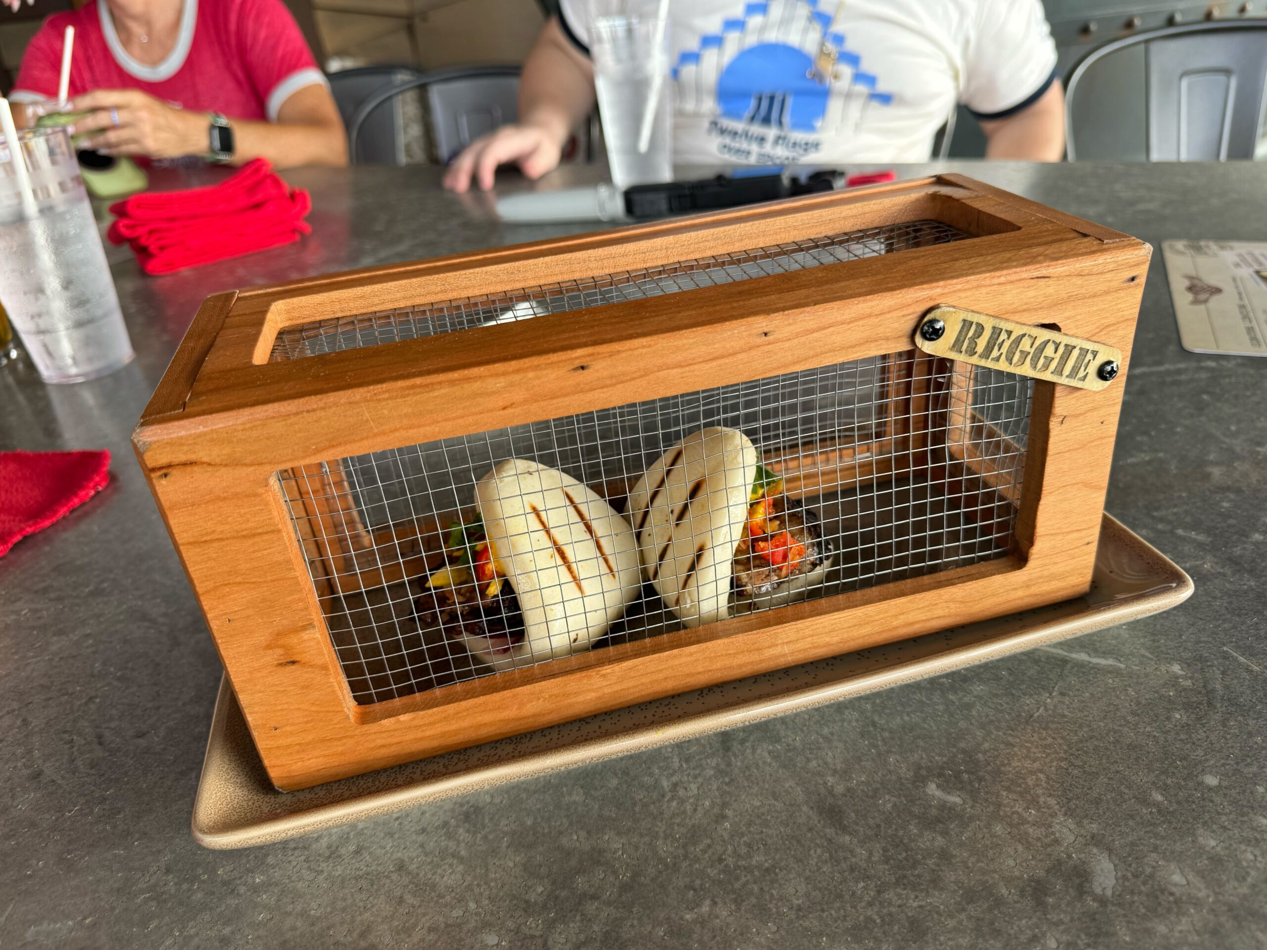 A wooden box with a mesh front contains two bao buns with delicious fillings inside. The box, labeled "REGGIT," is placed on a rectangular plate on a grey table, evoking the adventurous spirit of Jock Lindsey's Hangar Bar in Disney Springs.