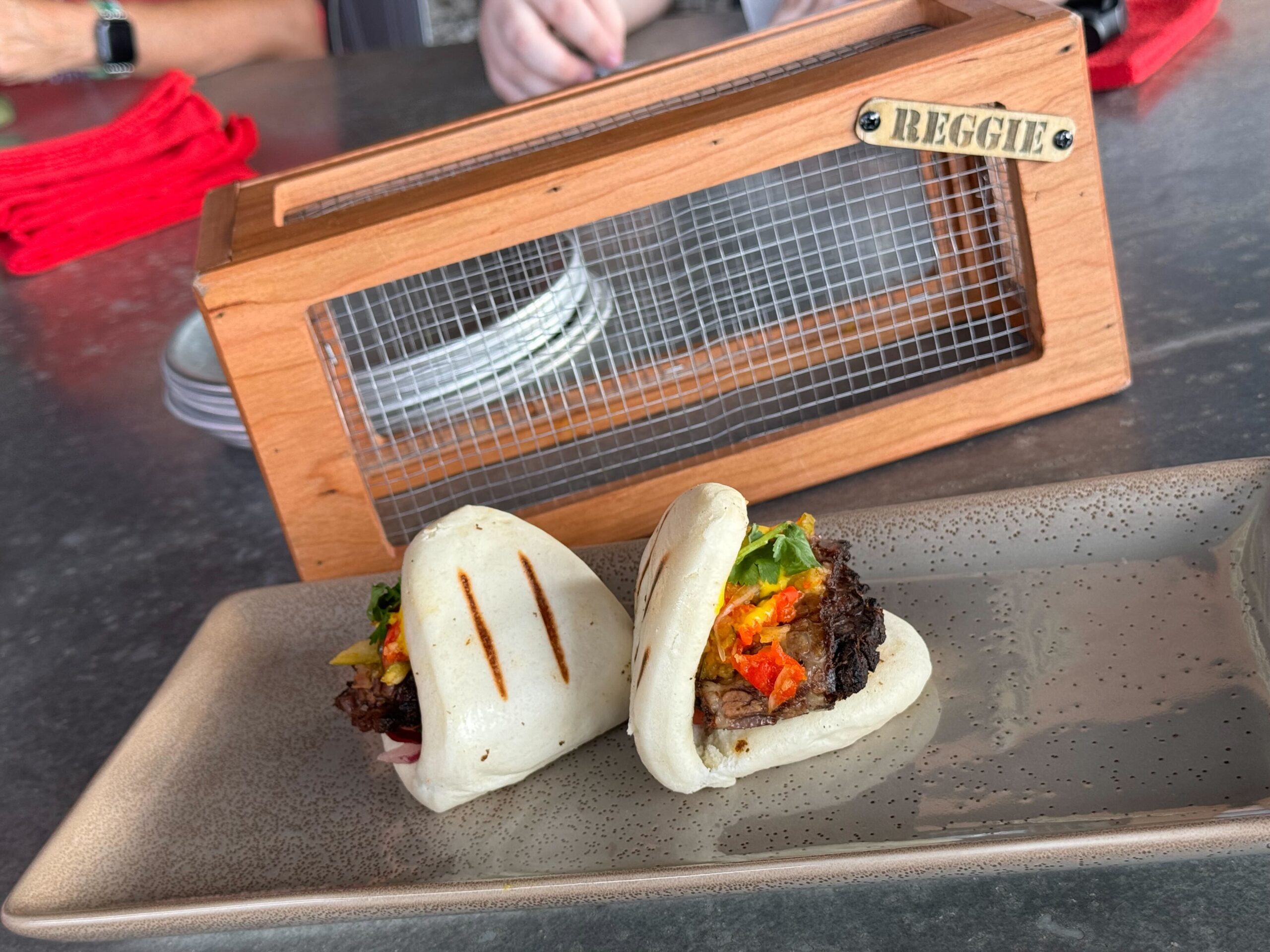 Two steamed bao buns filled with meat and vegetables on a rectangular plate, placed on a table at Jock Lindsey's Hangar Bar in Disney Springs. A wooden box labeled "Reggie" stands proudly in the background.