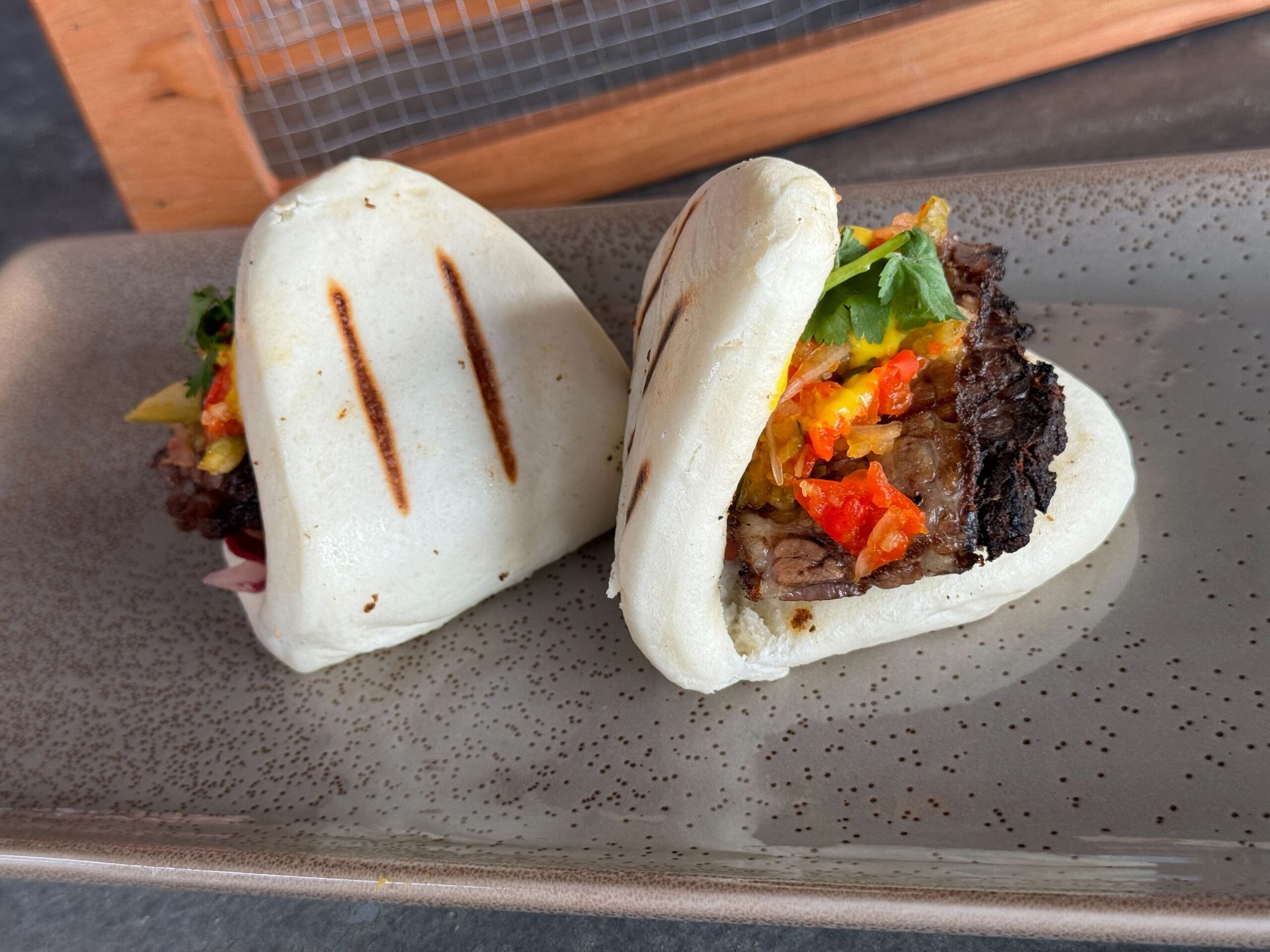 Two steamed buns filled with grilled meat, colorful pickled vegetables, and fresh herbs are served on a speckled rectangular plate at Jock Lindsey's Hangar Bar in Disney Springs.