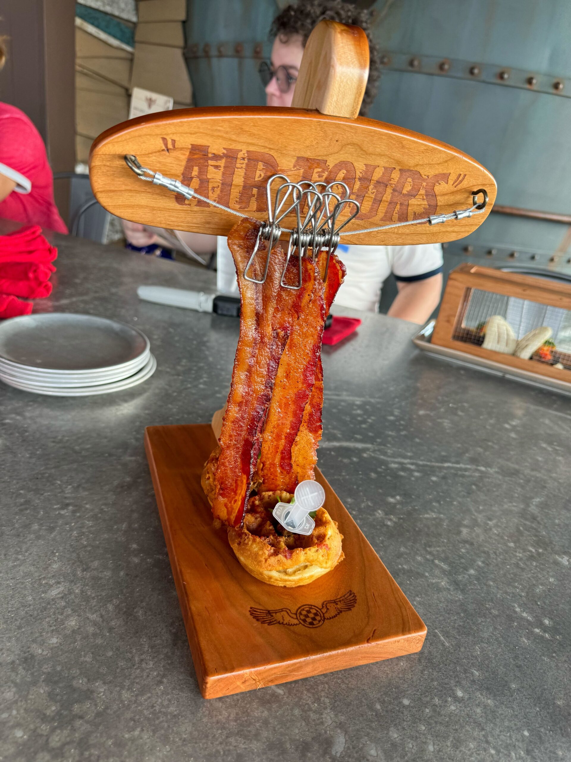 A decorative wooden biplane-shaped holder at Jock Lindsey's Hangar Bar in Disney Springs presents strips of crispy bacon hanging by clips, with a small serving of waffle and syrup on a wooden base.