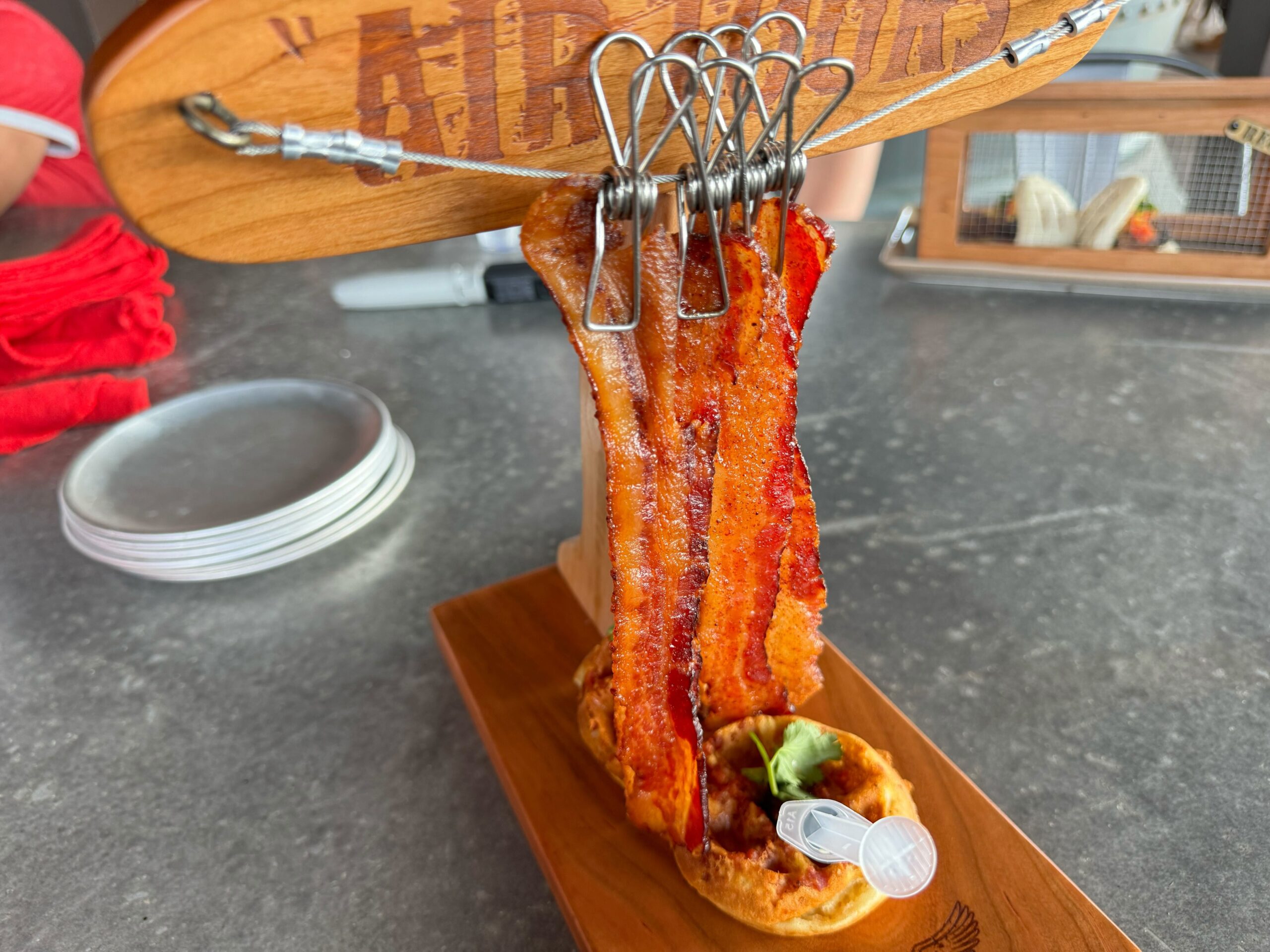 At Jock Lindsey's Hangar Bar in Disney Springs, a wooden board showcases crispy bacon strips suspended by metal clips above a small waffle, accompanied by a plastic pipette containing syrup.