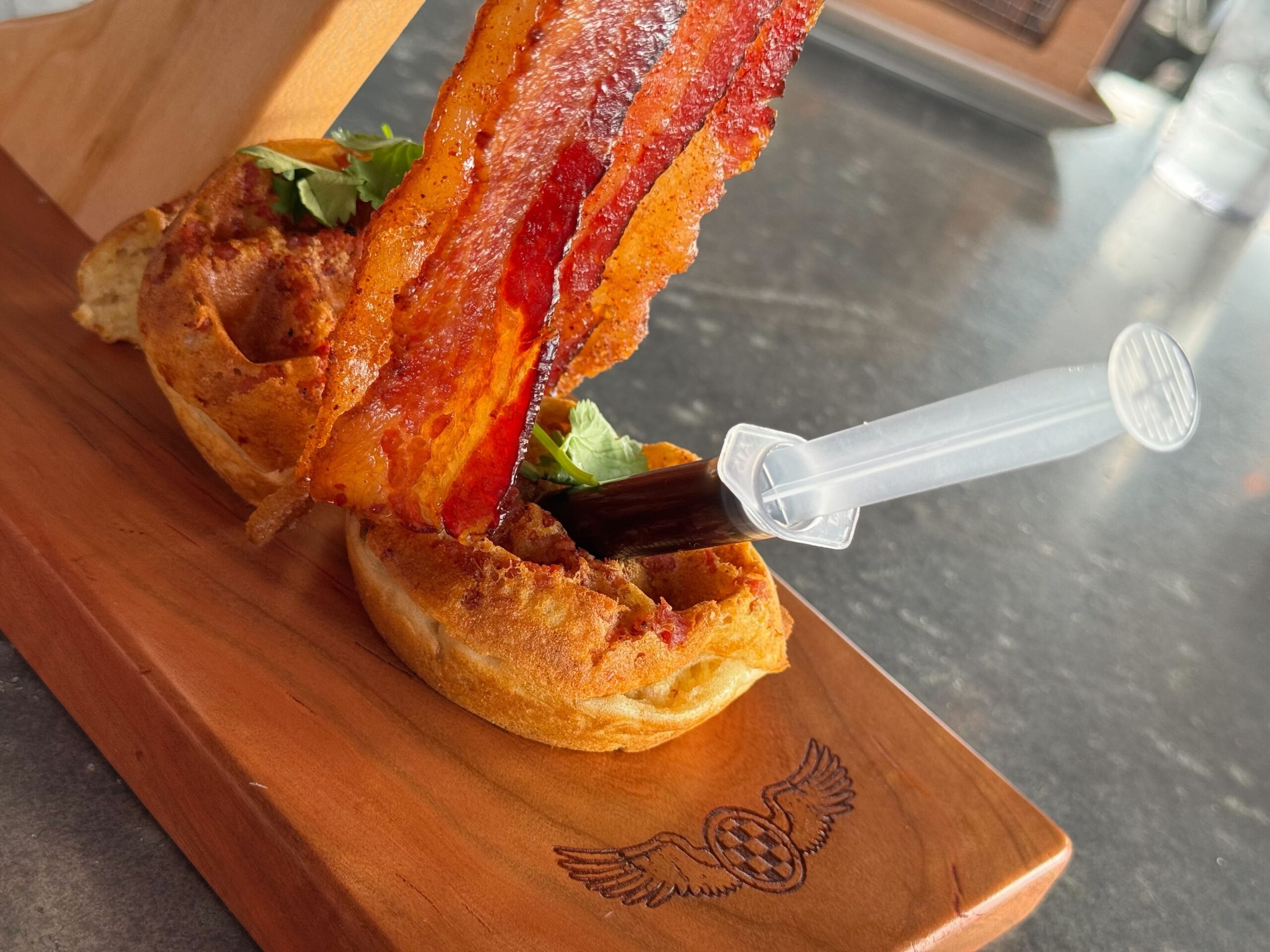 At Jock Lindsey's Hangar Bar in Disney Springs, a serving of waffles topped with bacon slices and a garnish, accompanied by a sauce-filled syringe, is placed on a wooden board.