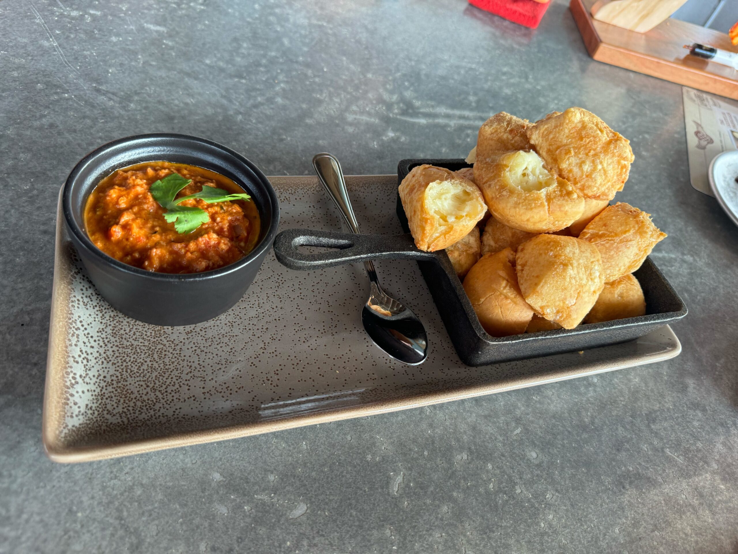 A dish with a bowl of red sauce garnished with herbs and a serving of fried bread rolls on a rectangular ceramic plate, accompanied by a spoon. This savory delight can be savored at Jock Lindsey's Hangar Bar in Disney Springs.