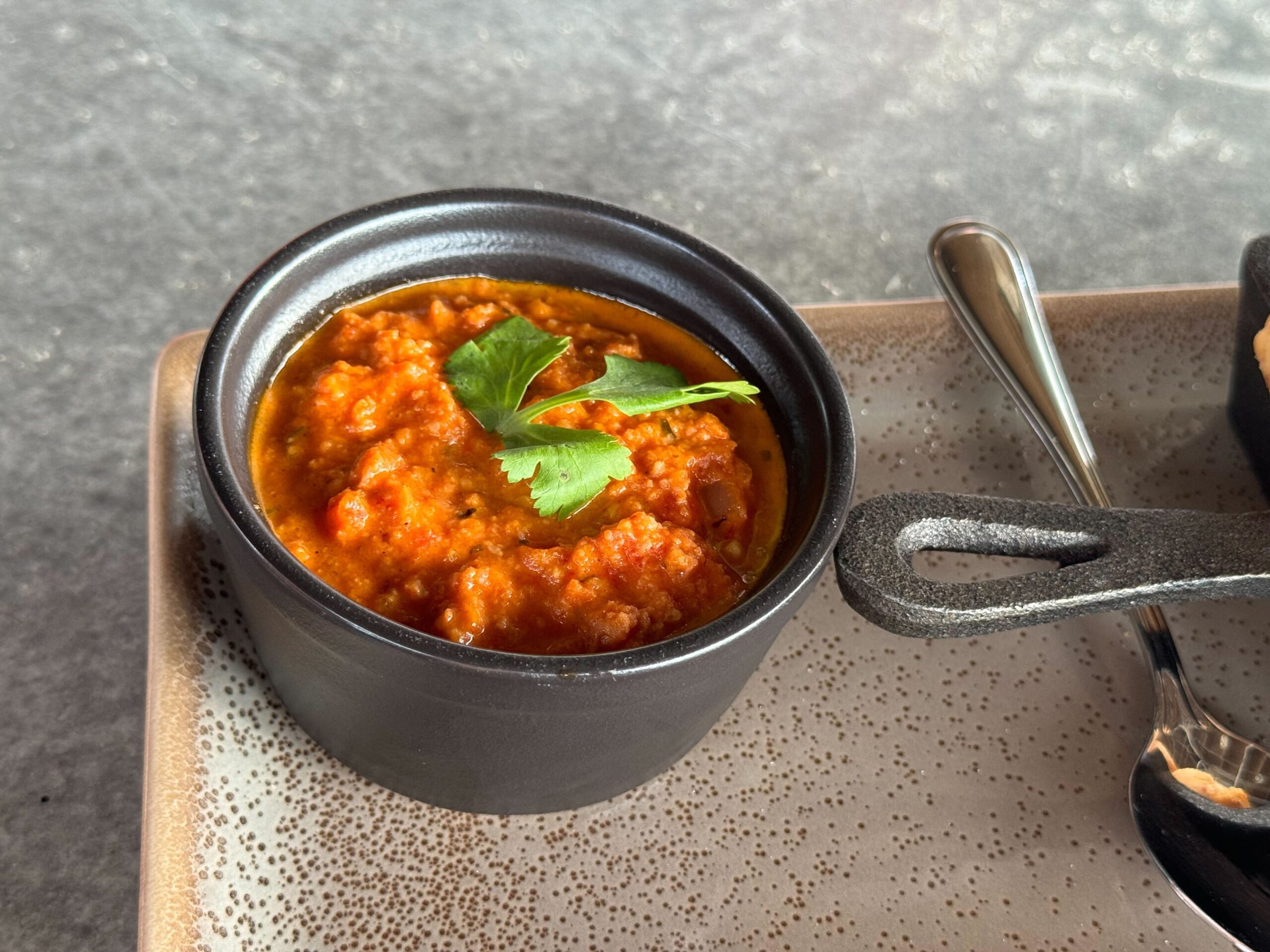 A small black bowl filled with red salsa garnished with a cilantro leaf, placed next to a metal spoon on a beige speckled tray, reminiscent of the eclectic vibes at Jock Lindsey's Hangar Bar in Disney Springs.