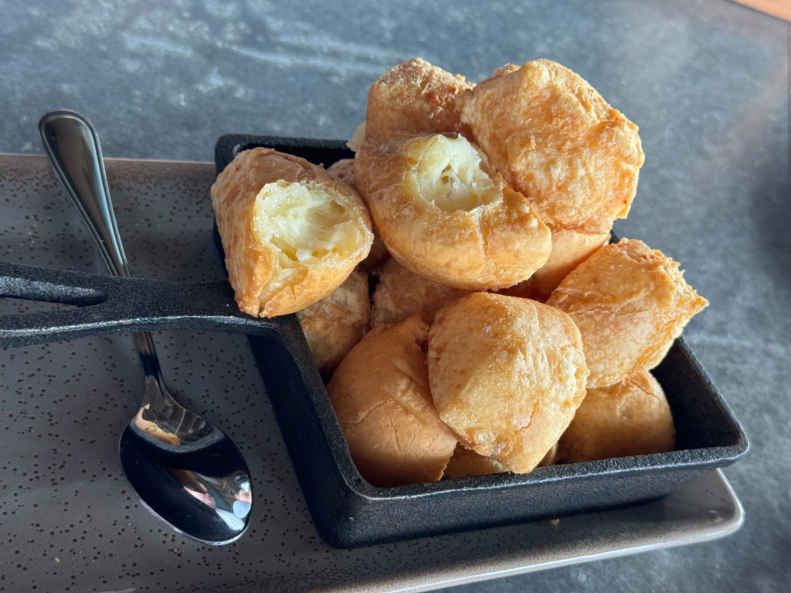 A black rectangular dish at Jock Lindsey's Hangar Bar in Disney Springs contains several pieces of fried dough, with one piece broken open to reveal a light, airy texture. A small metal spoon rests beside the dish on a speckled tray.