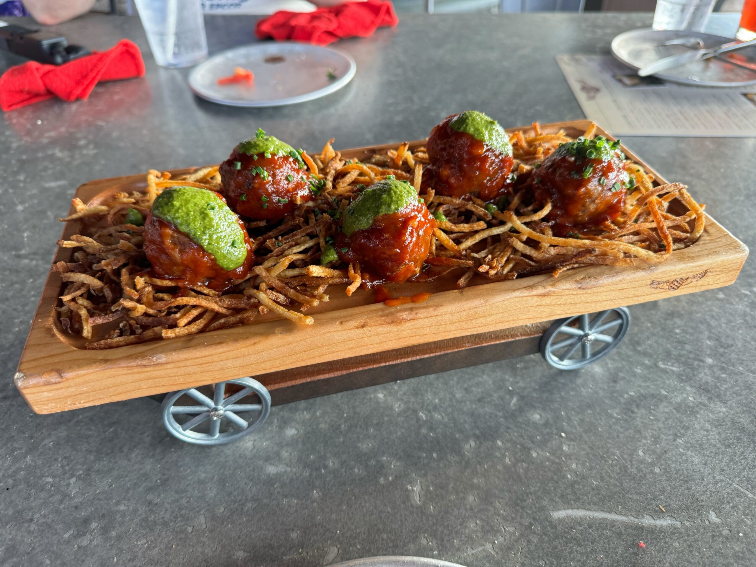 A wooden serving board holds six meatballs topped with green sauce, placed on a bed of crispy fries, presented on a small cart with wheels at Jock Lindsey's Hangar Bar in Disney Springs.