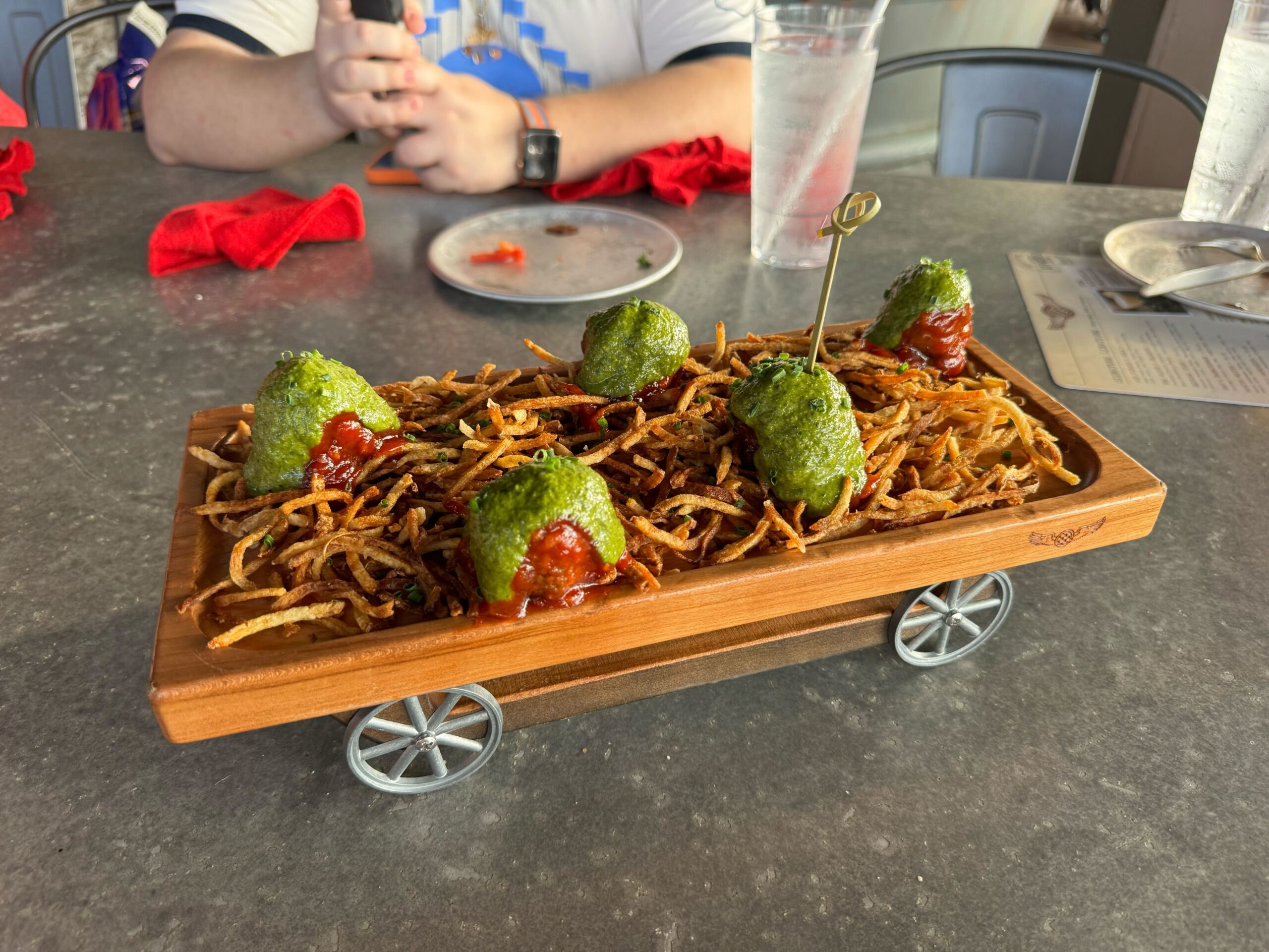 A wooden tray on wheels holds five green, ball-shaped food items topped with sauce, placed on a bed of fried strings at Jock Lindsey's Hangar Bar in Disney Springs. A person is seated in the background at a table.