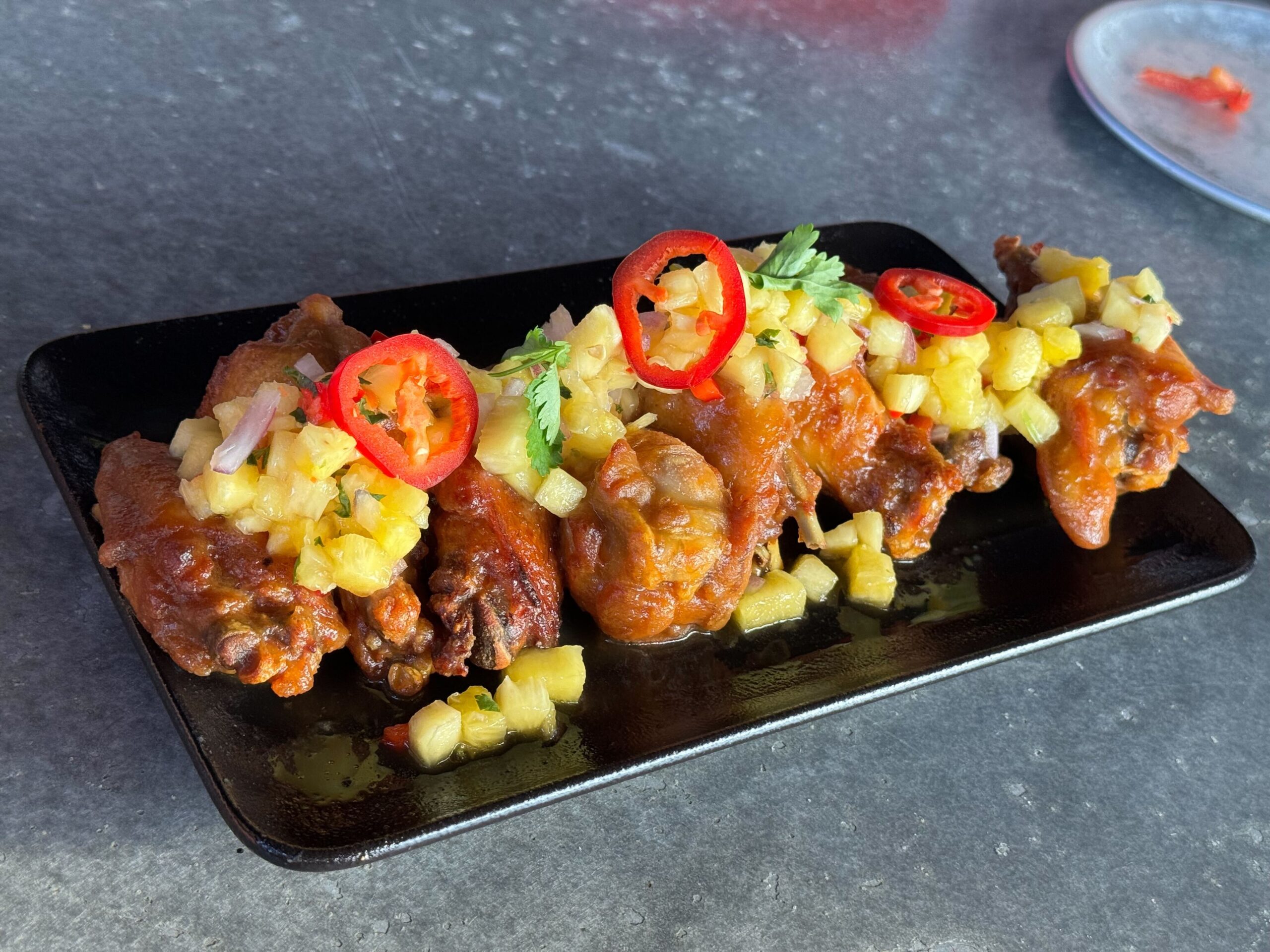 Chicken wings garnished with chopped pineapple, red chili slices, and cilantro on a black rectangular plate at Jock Lindsey's Hangar Bar in Disney Springs.