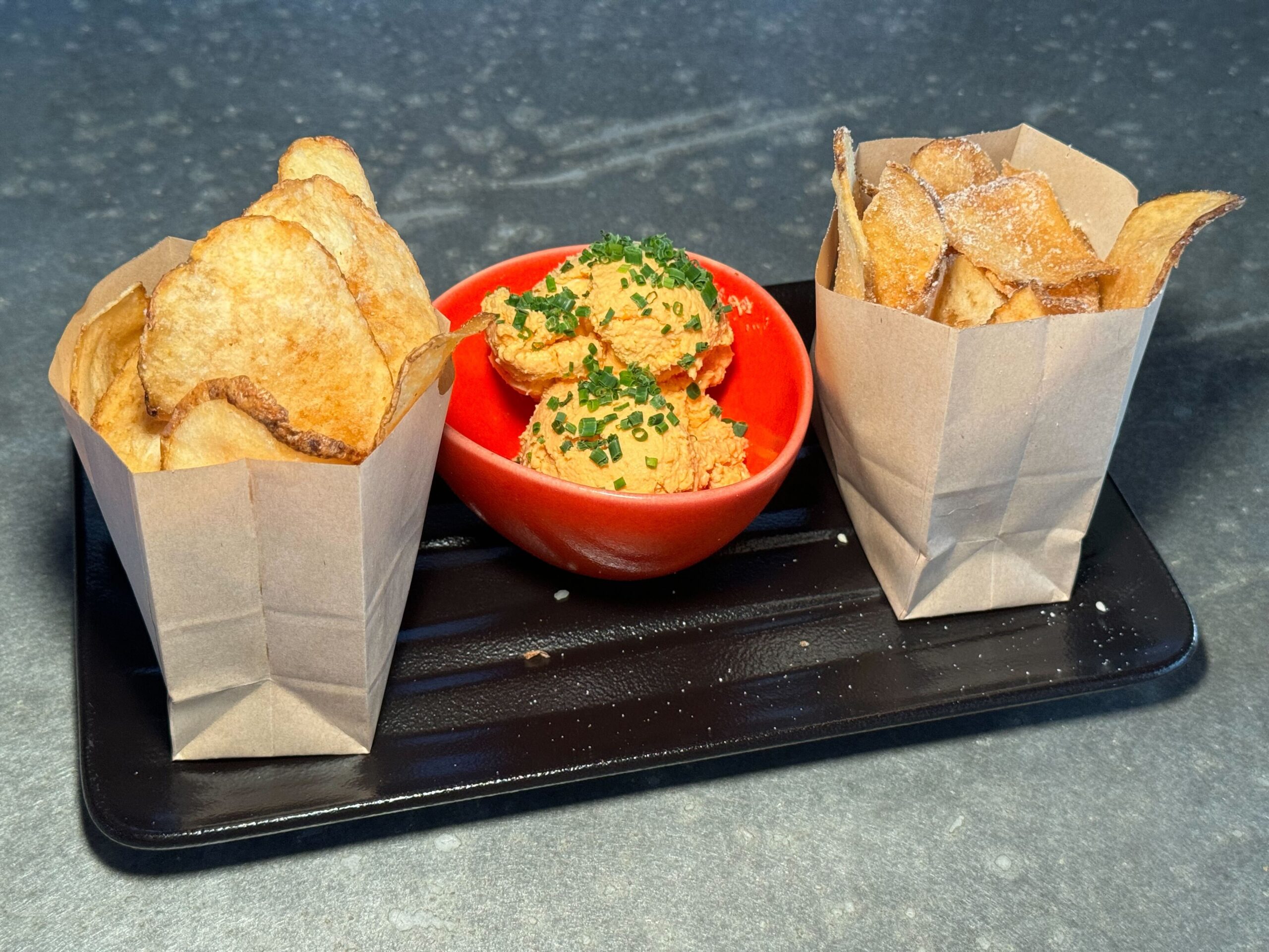 Paper bags filled with homemade potato chips, accompanied by a red bowl of chive-topped dip, are served on a black rectangular tray at Jock Lindsey's Hangar Bar in Disney Springs.
