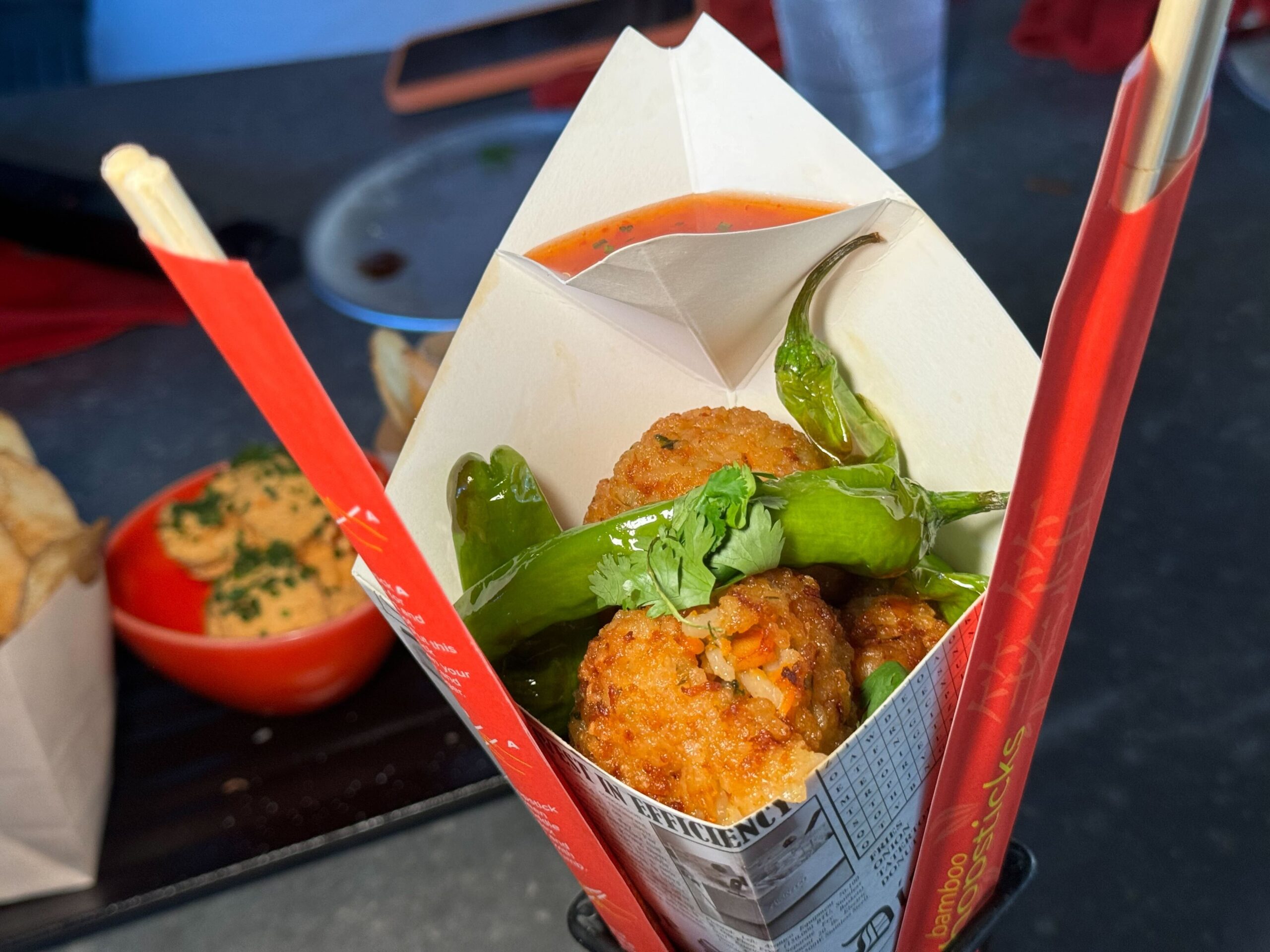 Close-up of a takeout container holding breaded fried bites garnished with green peppers and cilantro, with a red dipping sauce cup in the background. Perfect for your next meal at Jock Lindsey's Hangar Bar in Disney Springs.