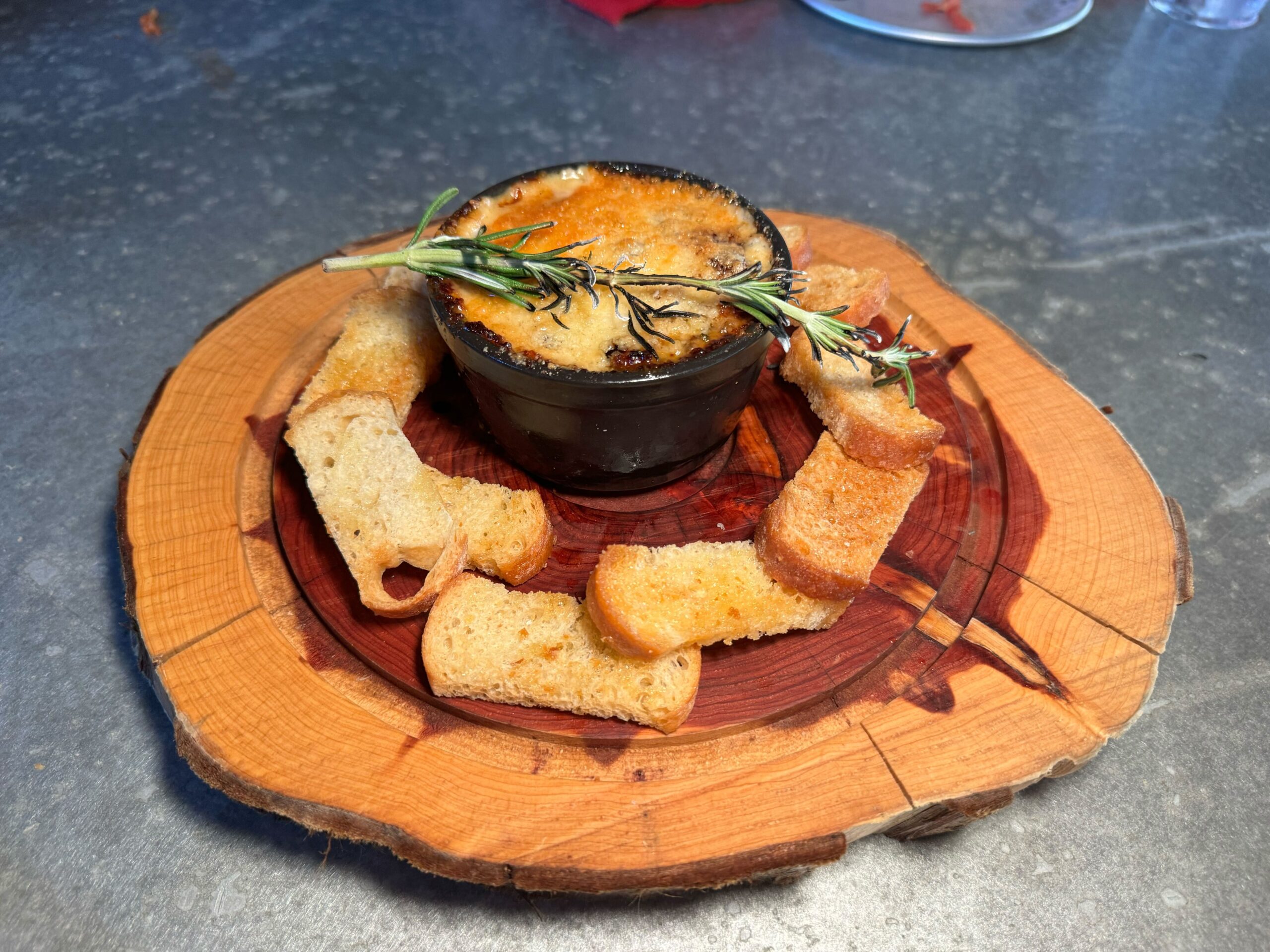A black bowl with a cheese-topped dip garnished with rosemary, surrounded by toasted bread slices, is placed on a round wooden serving board at Jock Lindsey's Hangar Bar in Disney Springs.