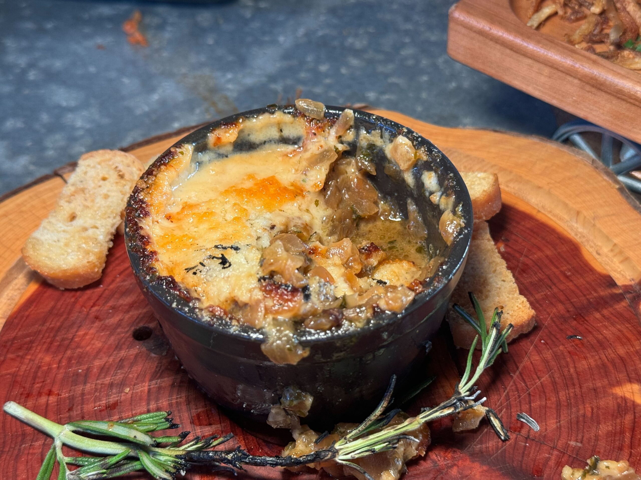 A small black dish of baked onion soup with melted cheese on top, served with toasted baguette slices and garnished with a sprig of rosemary graces the wooden board at Jock Lindsey's Hangar Bar in Disney Springs.