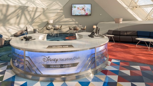 Disney Vacation Club member lounge with a reception desk, “Welcome Home” sign, modern seating, and a TV screen in a brightly lit room with geometric patterned carpeting.