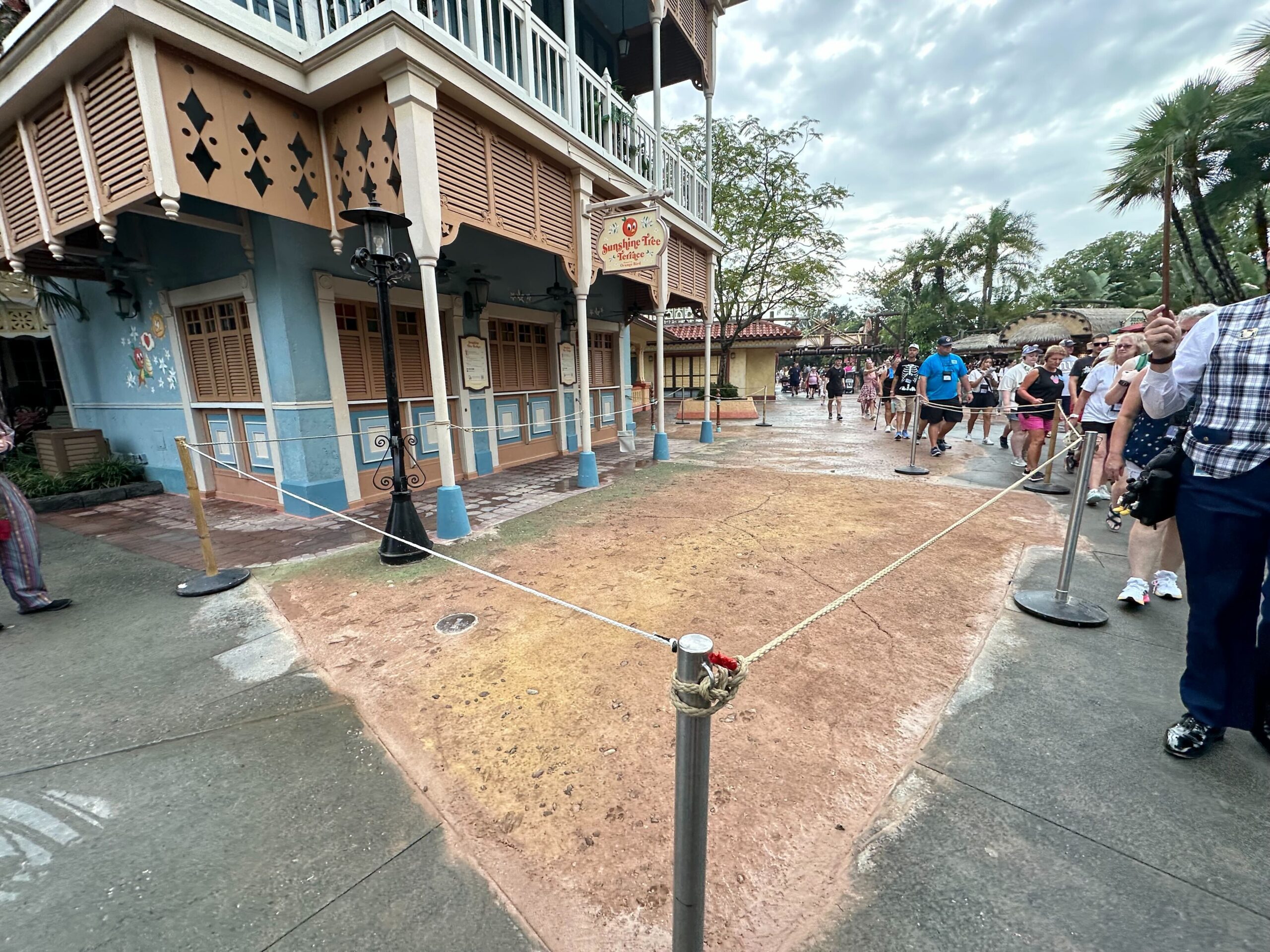 Empty roped-off area on a street corner near a building with a blue facade, with people standing nearby on a cloudy day.