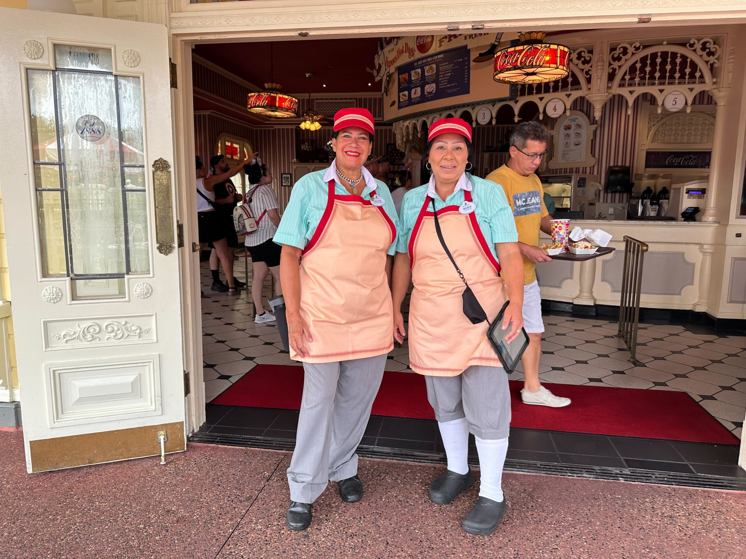 Two employees wearing uniforms, standing by the entrance of a store or restaurant with a customer exiting in the background carrying a tray.