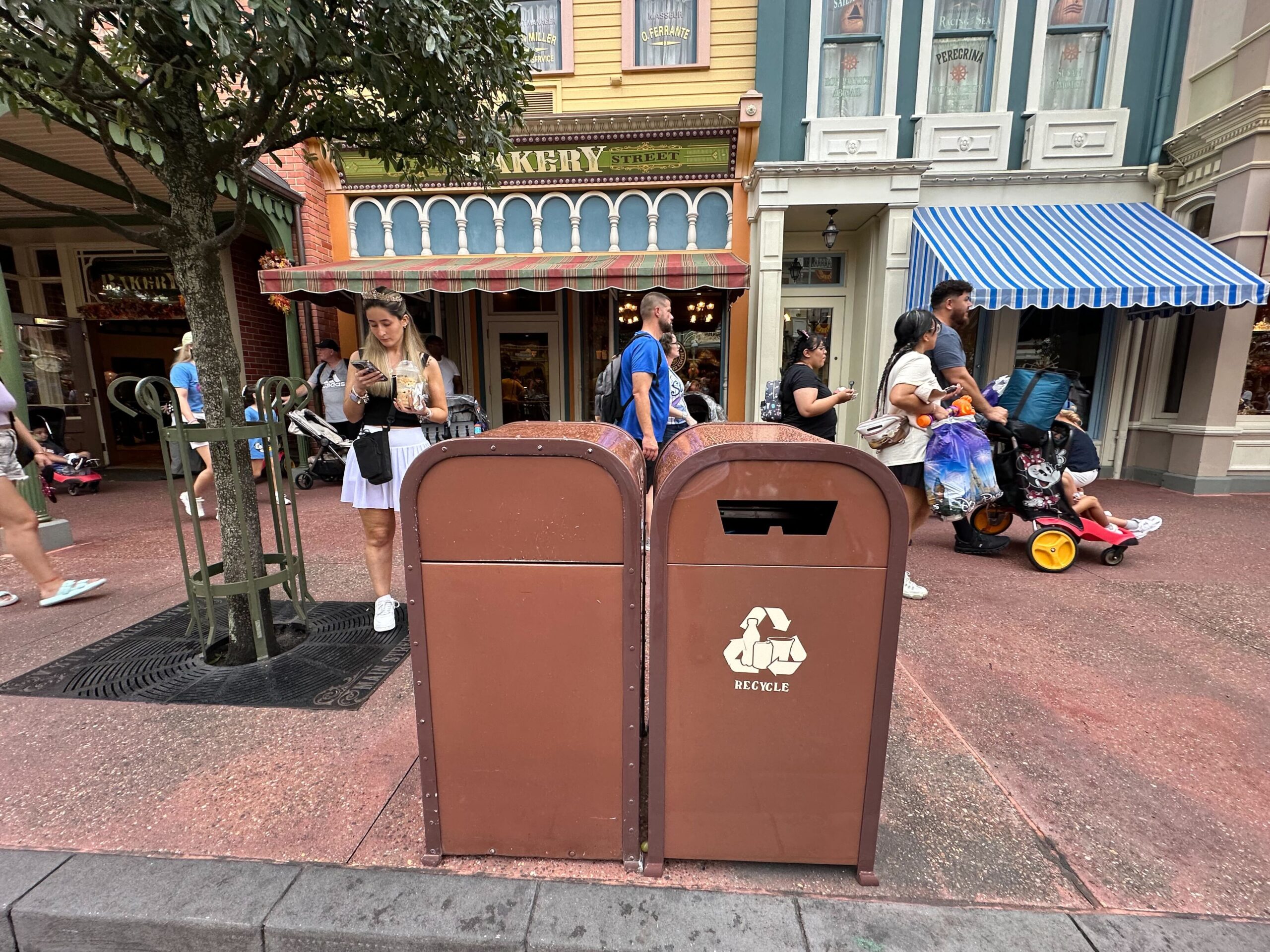 A brown recycling bin and a trash can are placed side by side on a busy sidewalk in front of a bakery. People are walking and sitting in the background.