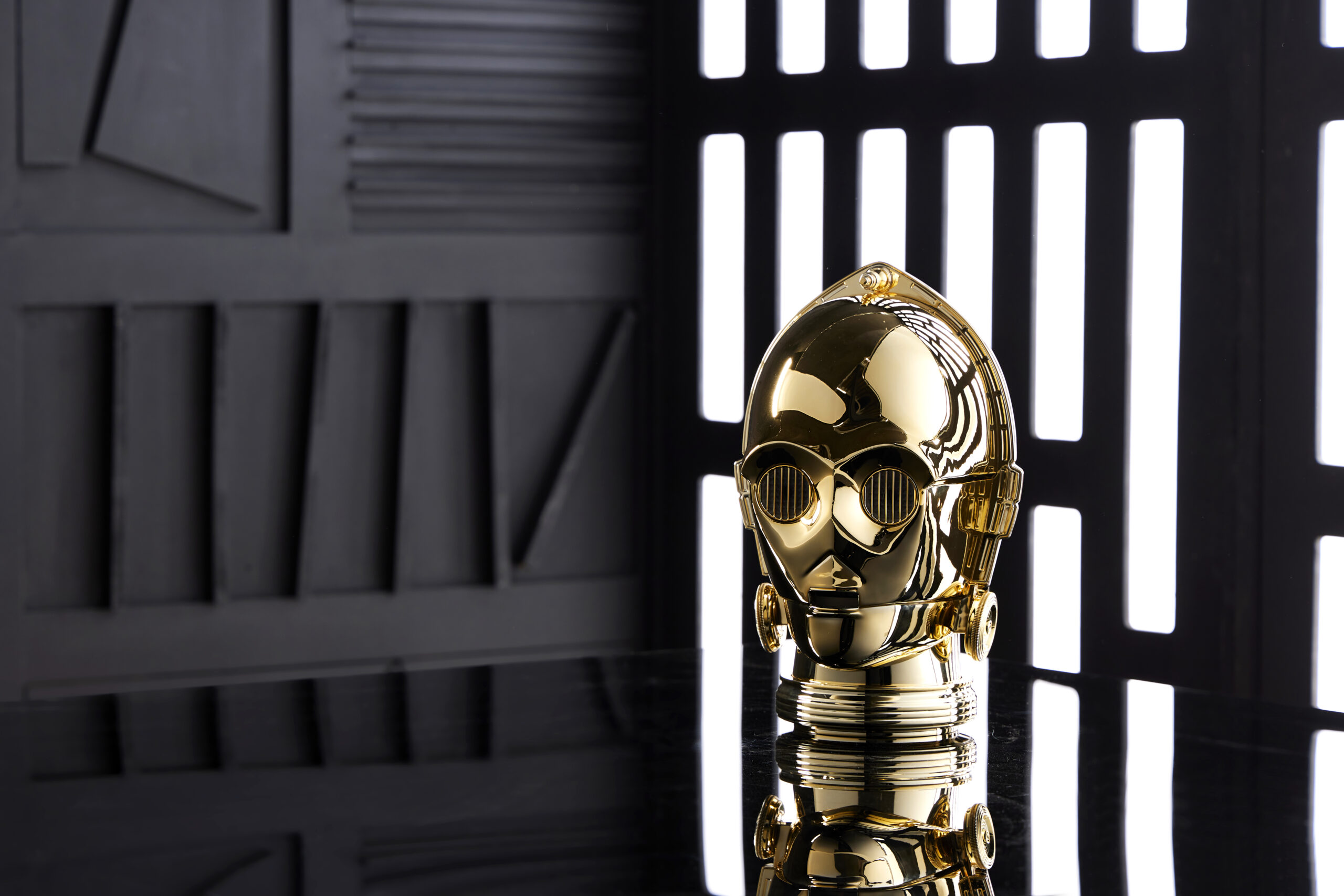 A gold, robot-like head stands on a reflective surface against a black and white striped backdrop.