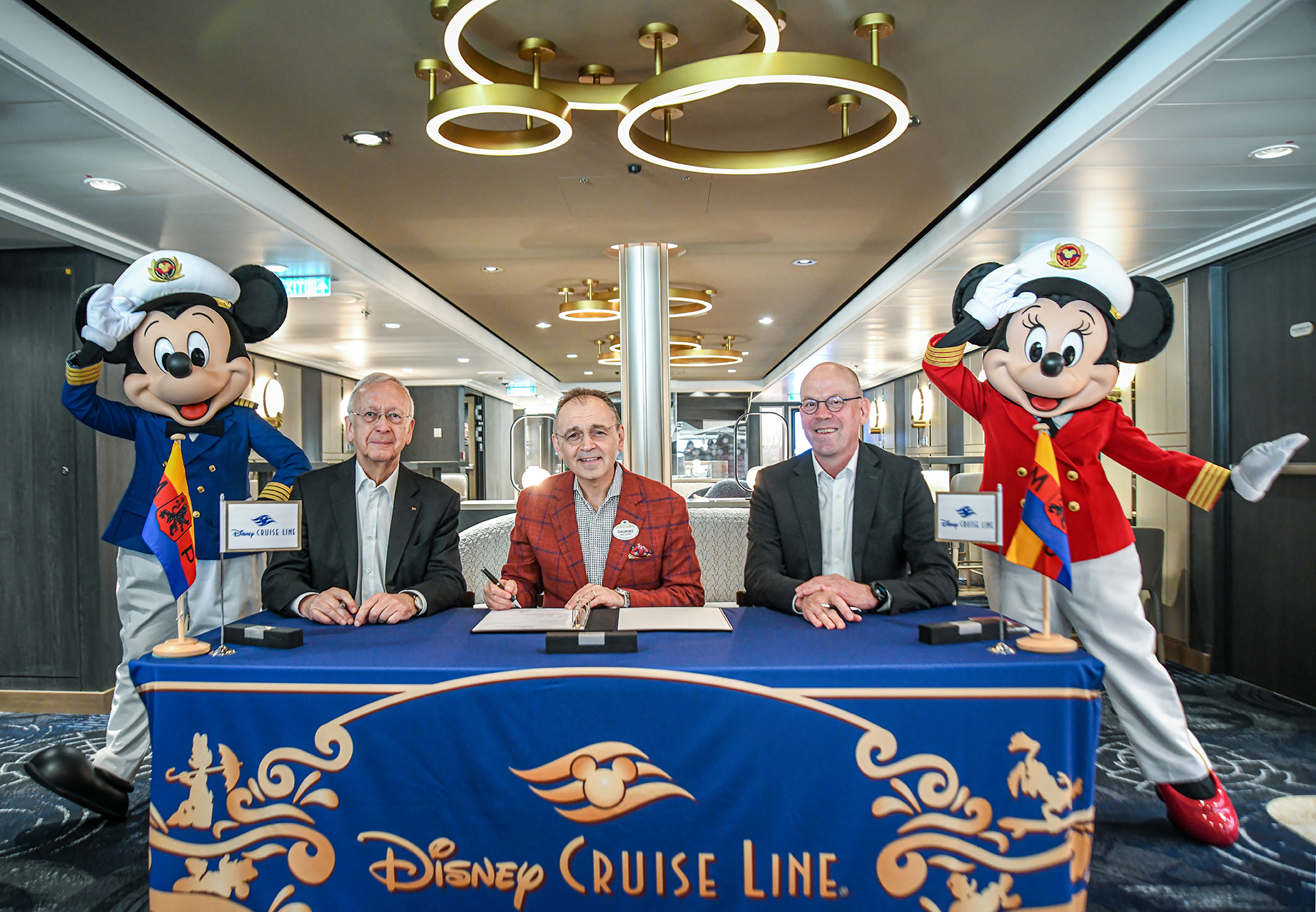 Three men sit at a table with Disney Cruise Line branding. Two mascots dressed as captains stand beside them.