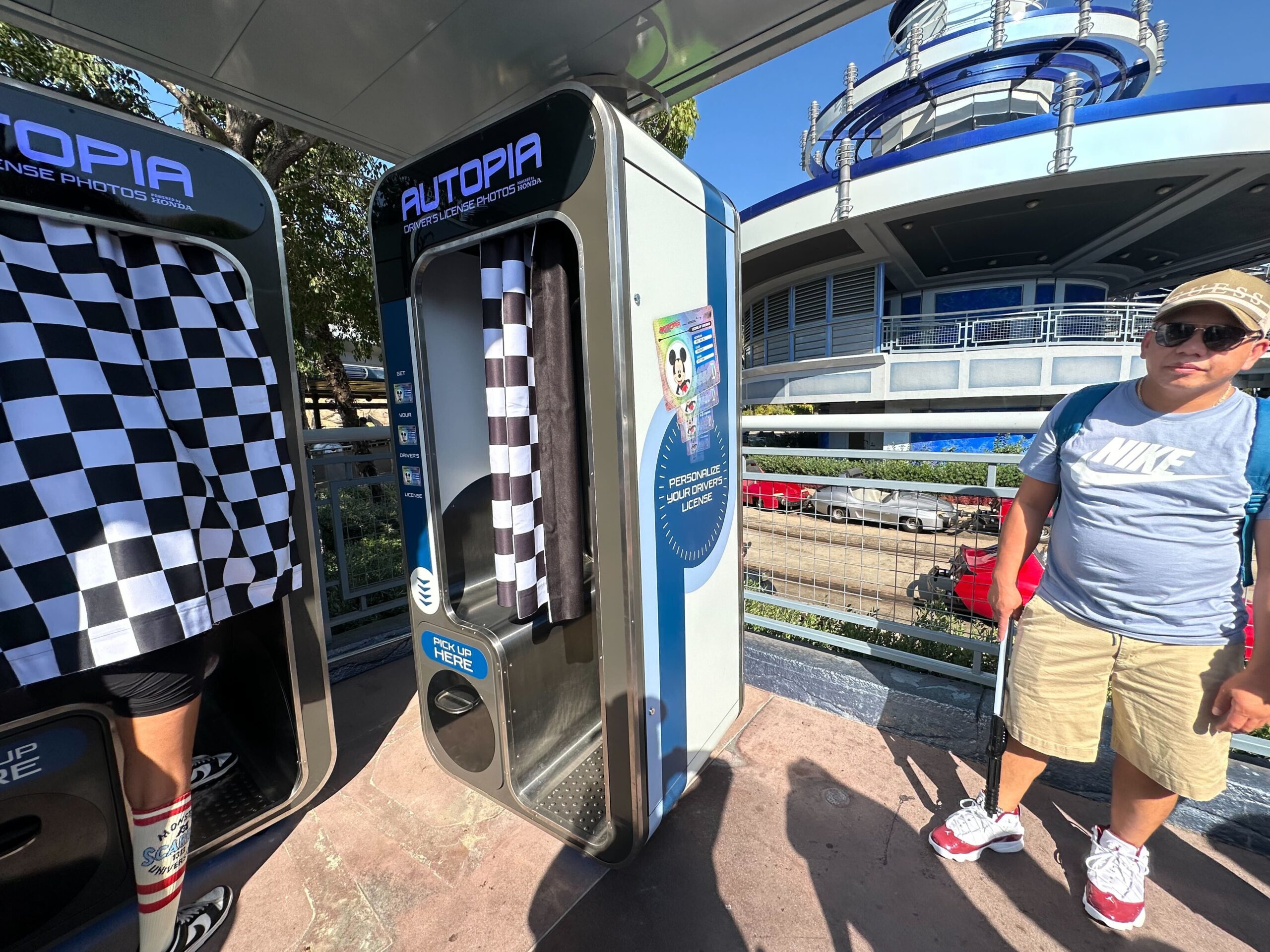 A person standing next to an Autopia ride photo booth holding souvenir photos, with another person interacting. Steps and a futuristic building are visible in the background.