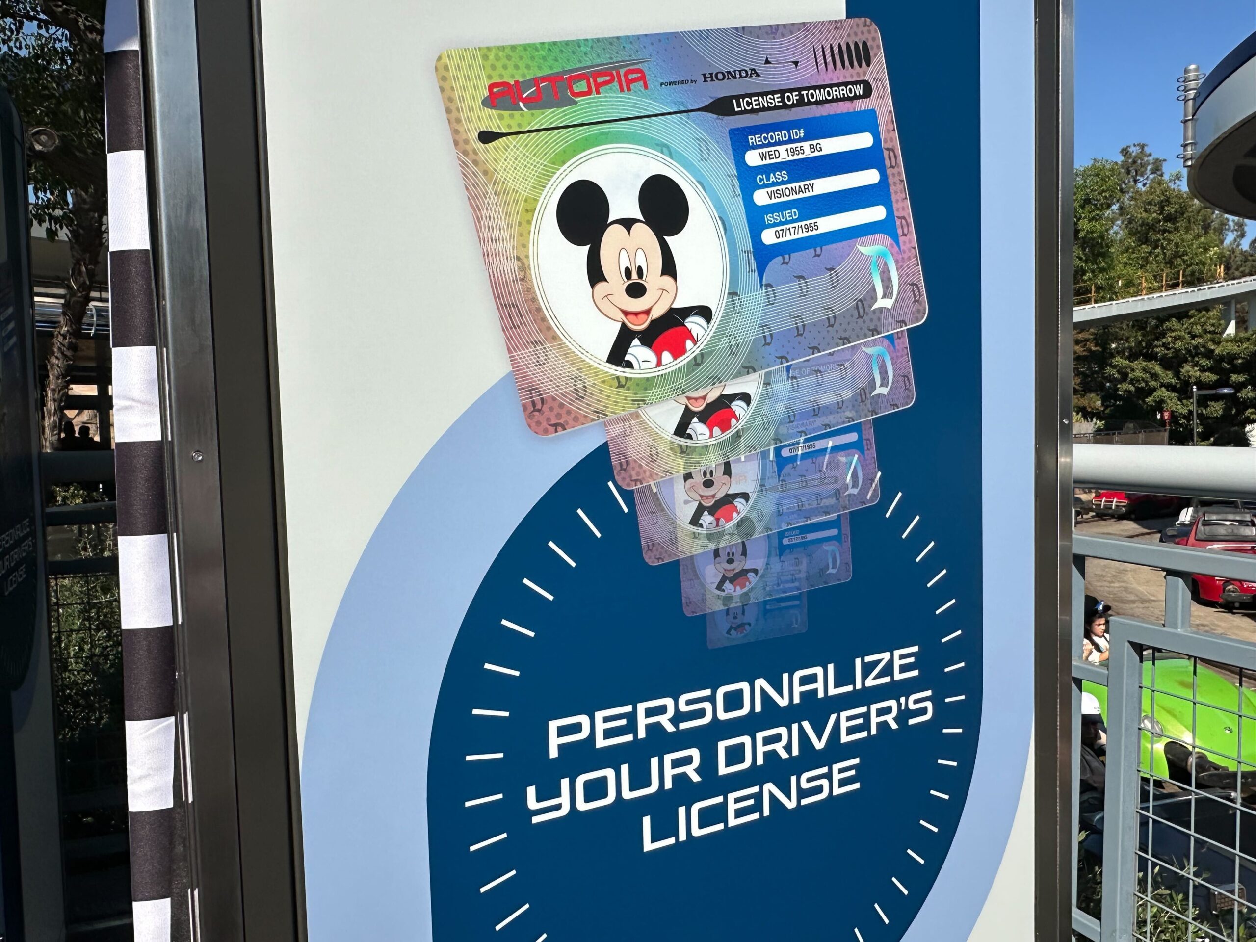 Sign advertising personalized driver's licenses featuring a cartoon character, displayed on a blue background with trees and sunlight in the background.