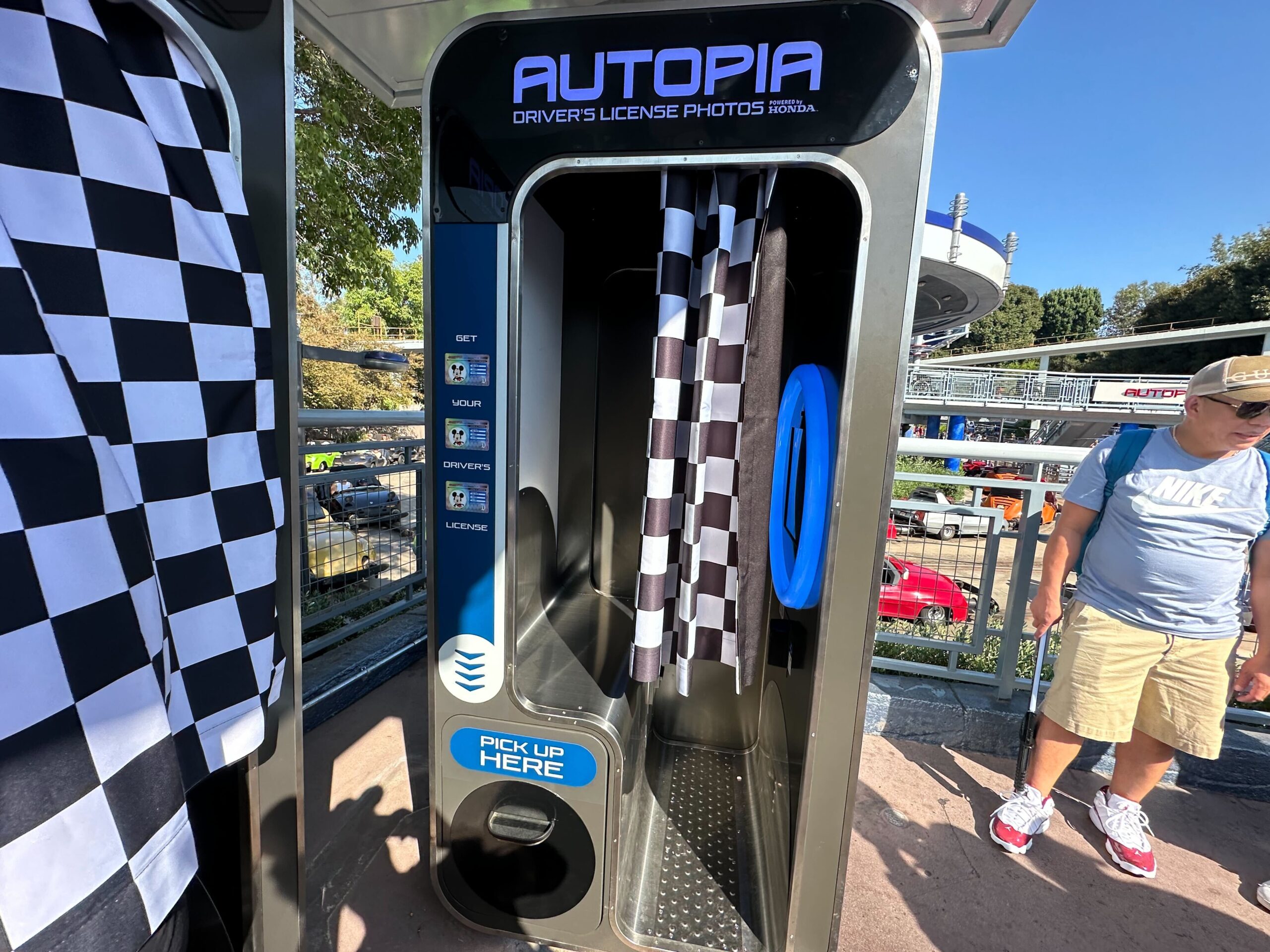Autopia Driver's License Photos booth with a checkered curtain and "Pick Up Here" sign, located outdoors on a sunny day. A person stands nearby.