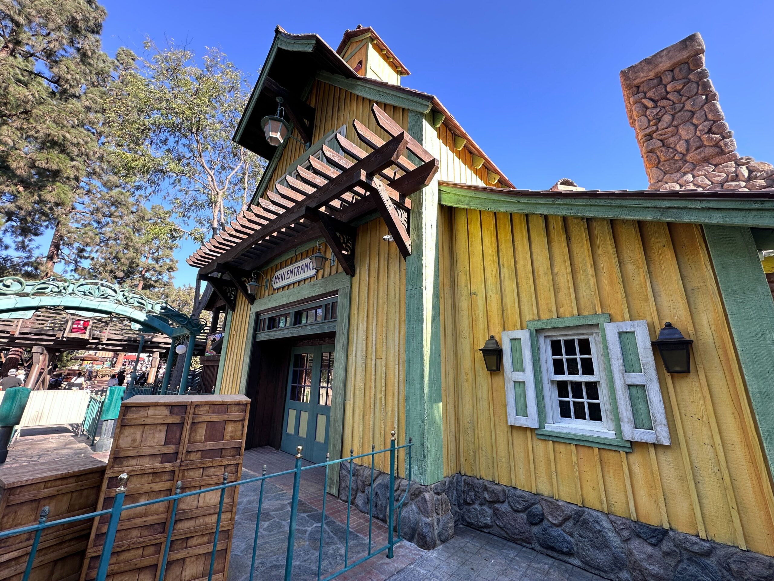 The rustic building, reminiscent of Tiana's Bayou Adventure at Disneyland, features a yellow wooden exterior with green accents and a stone chimney. Stacked crates and a gated path add charm to the scene, framed by trees against a brilliant blue sky.