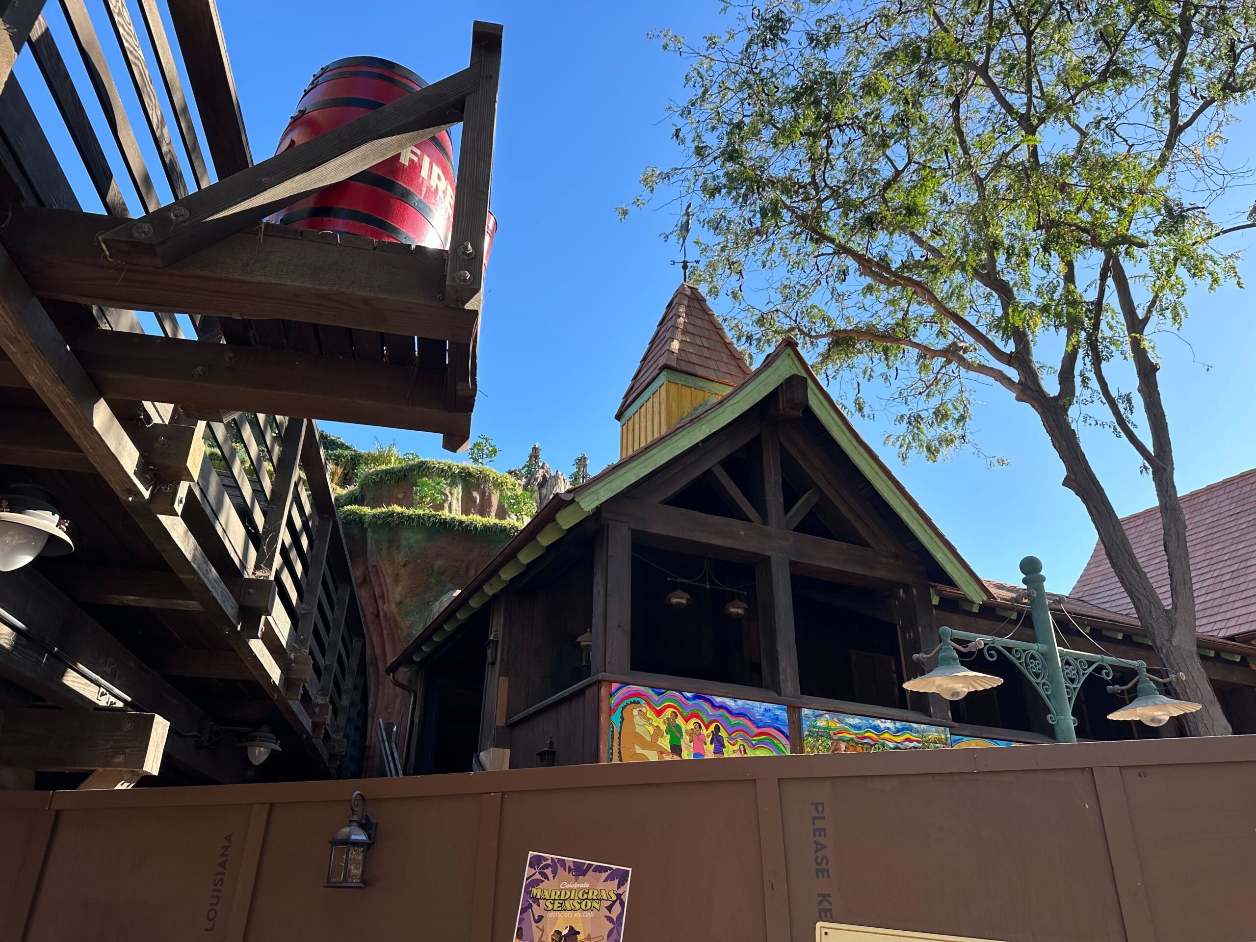 A rustic wooden building with a pointed roof stands behind a construction wall at Disneyland. Nearby, a large red barrel rests on an elevated platform, while a tree sways gently under the clear blue sky, hinting at the magic of Tiana's Bayou Adventure soon to come.