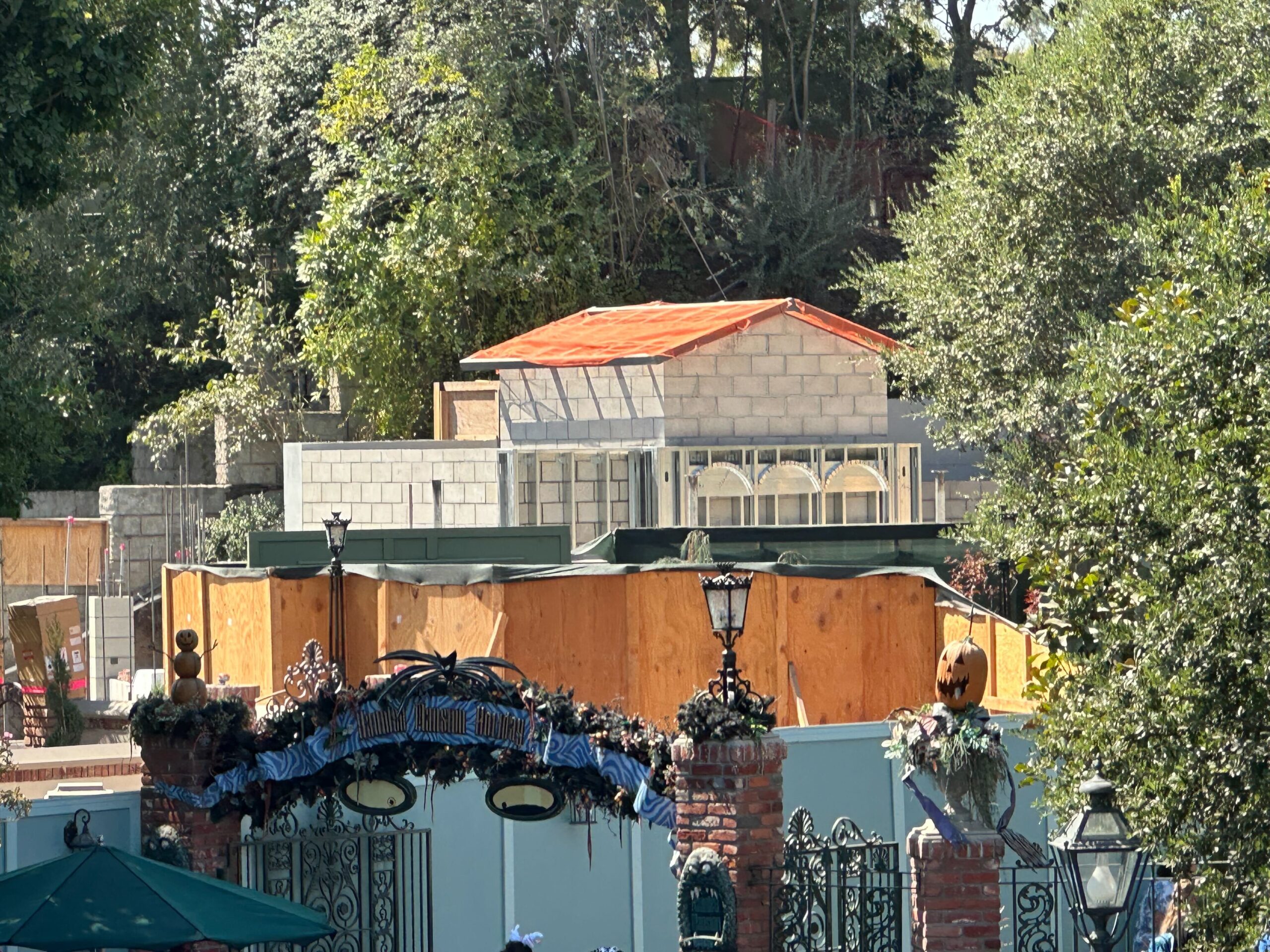 A construction site behind an open-space area with trees. The structure being built has concrete block walls and a roof partially covered with orange material. The surroundings include some scaffolding and wooden barriers.