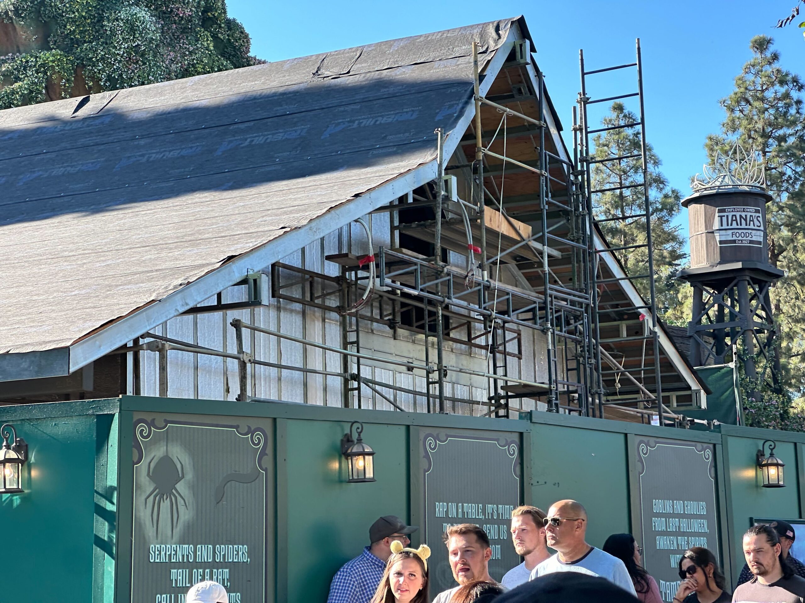 Scaffolding on a building under construction, surrounded by green construction barriers. A "Tiana's Foods" water tower is visible in the background. People are walking in front.