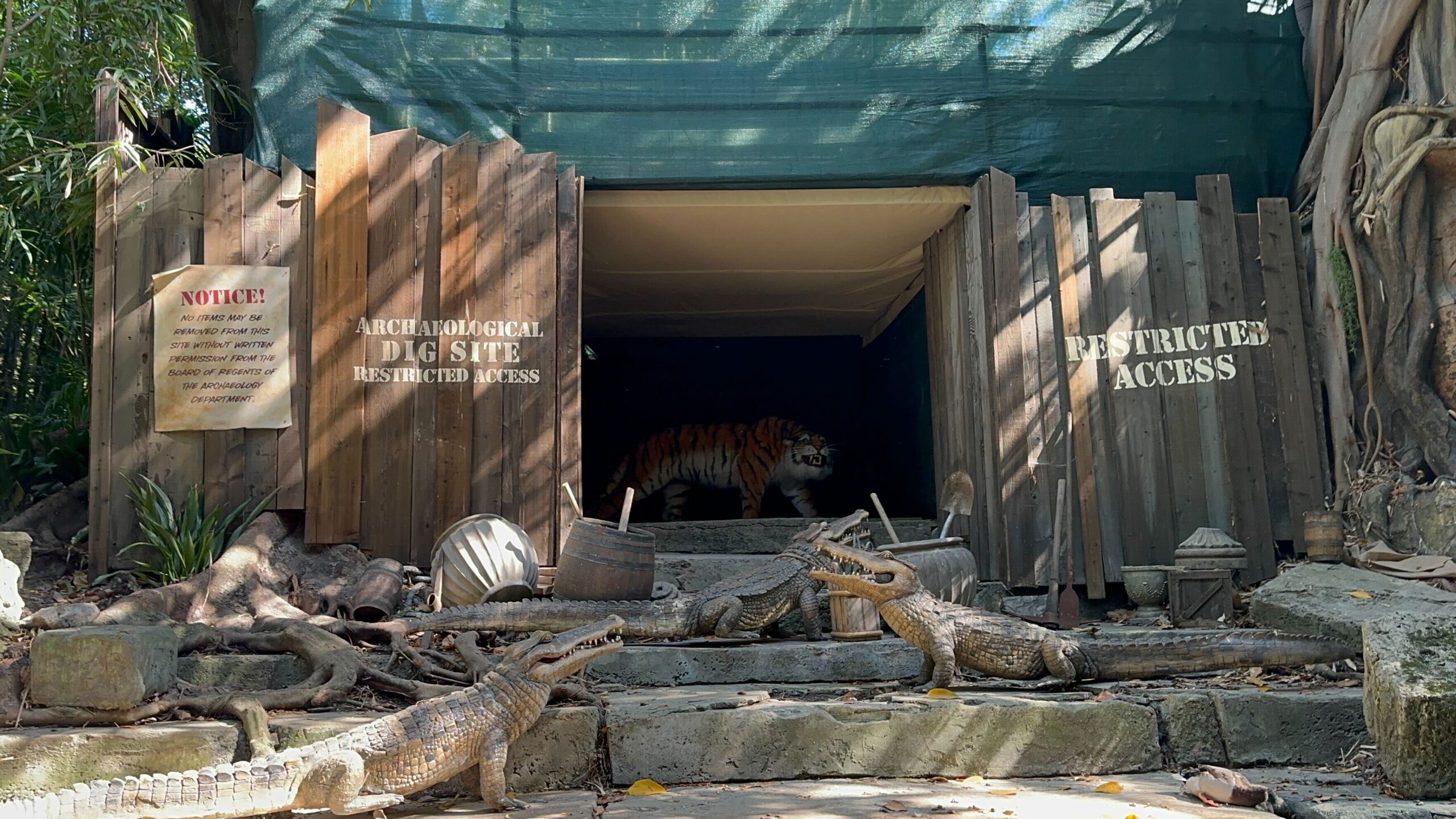 An exhibit featuring a tiger behind a wooden gate labeled "Archaeological Dig Site Restricted Access," with statues of crocodiles in the foreground.