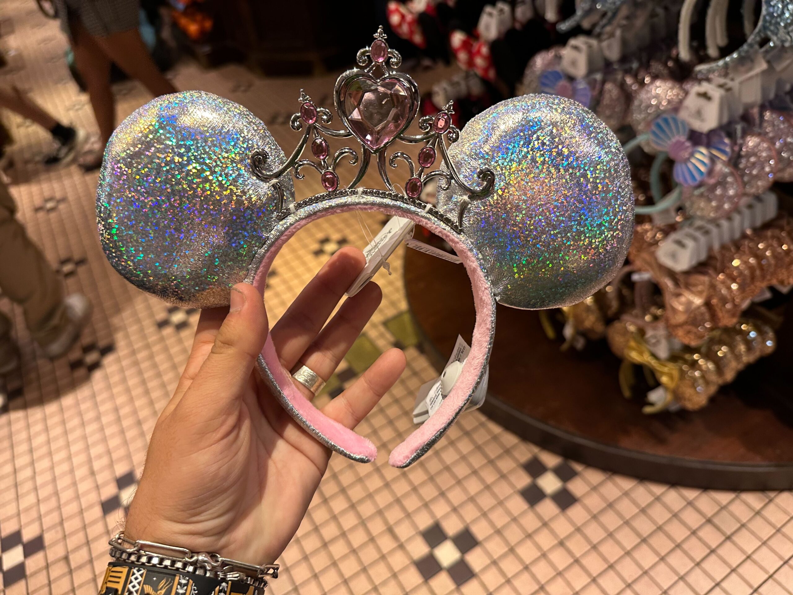 A hand holds a sparkly mouse ear headband adorned with a silver tiara and heart-shaped jewel, reminiscent of the whimsical charm found in Disn-inspired collections, displayed elegantly in a store with other enchanting headbands in the background.