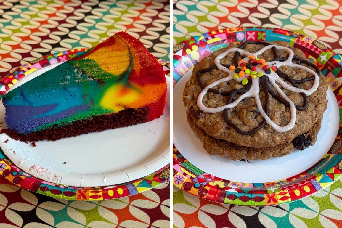 A slice of rainbow cake and a large decorated cookie with white icing and colorful sprinkles are placed on paper plates with colorful borders. The tablecloth beneath is patterned with overlapping ovals.