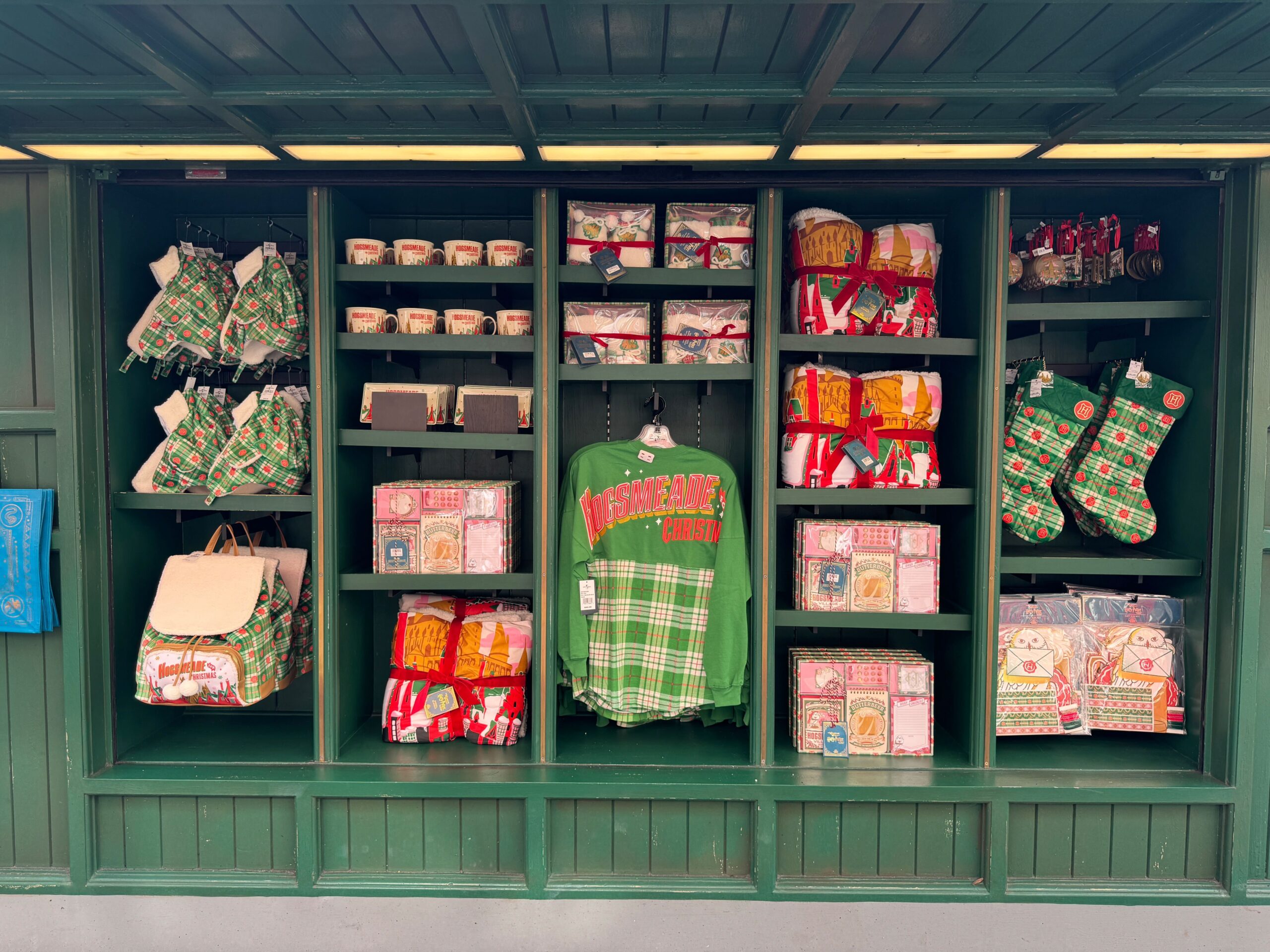 Display of holiday-themed merchandise in a green shelf unit, including mugs, shirts, kitchen items, stockings, and bags.