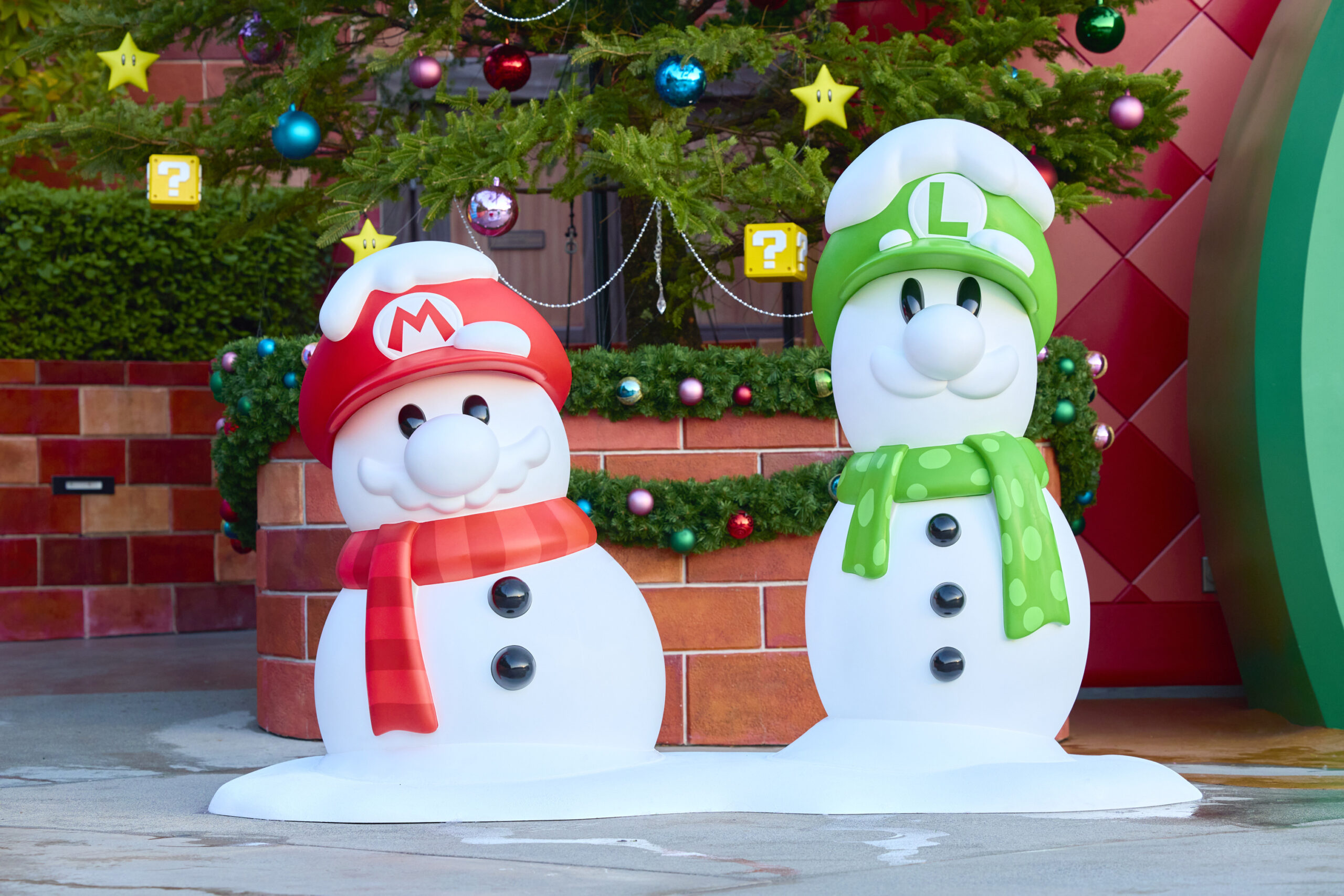 Two snowman figures wearing hats with letters "M" and "L" stand cheerfully in front of a decorated Christmas tree, adorned with baubles and yellow question mark cubes, celebrating the whimsical charm of Super Nintendo World during the festive season.