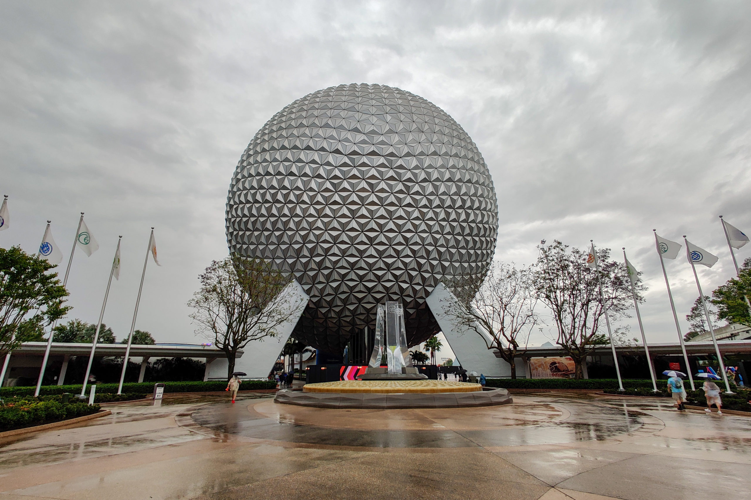 Spherical structure at Epcot with a geometric design, surrounded by flags and trees, under a cloudy sky.