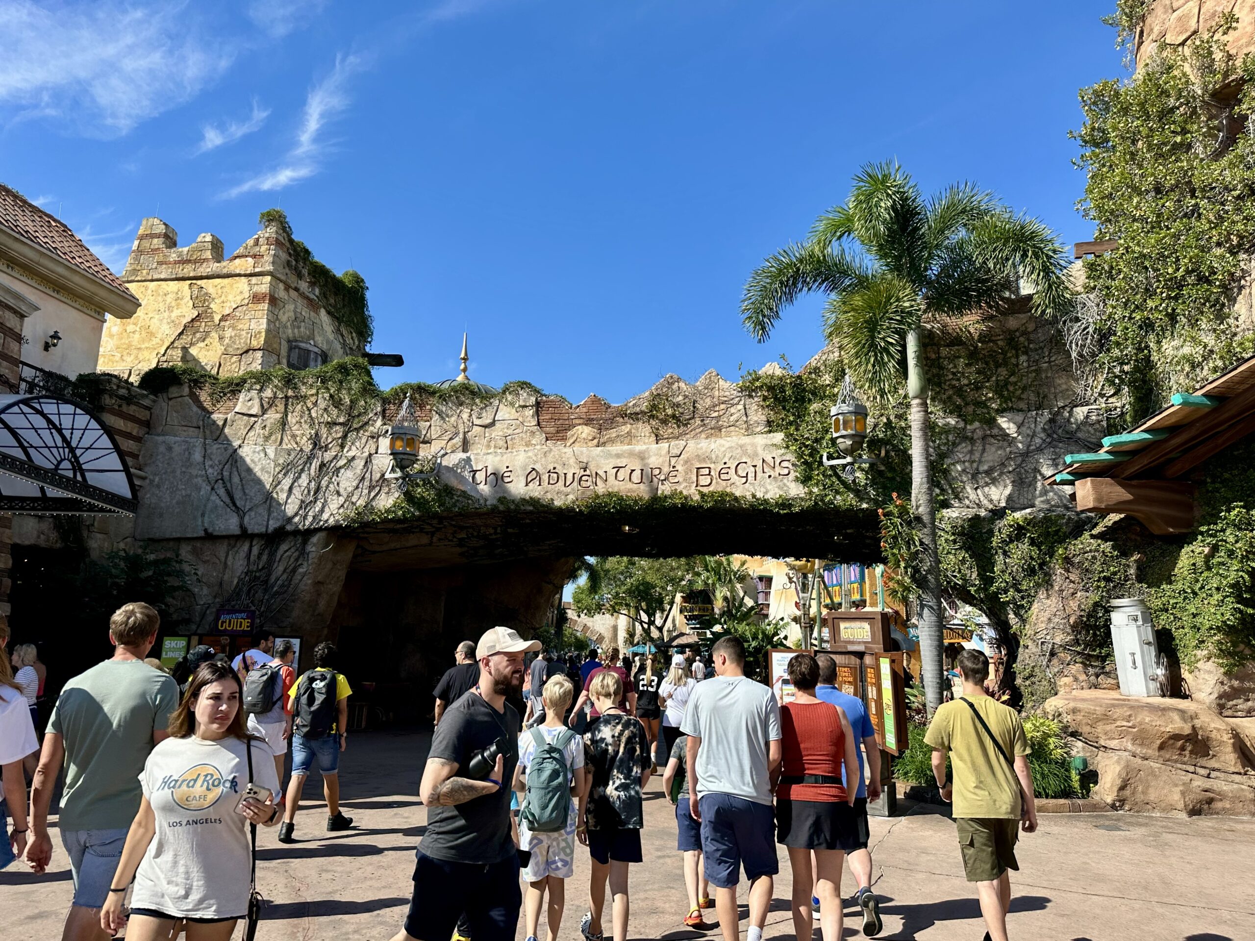 People walking towards an entrance marked "The Adventure Begins" surrounded by lush greenery and palm trees under a bright blue sky.