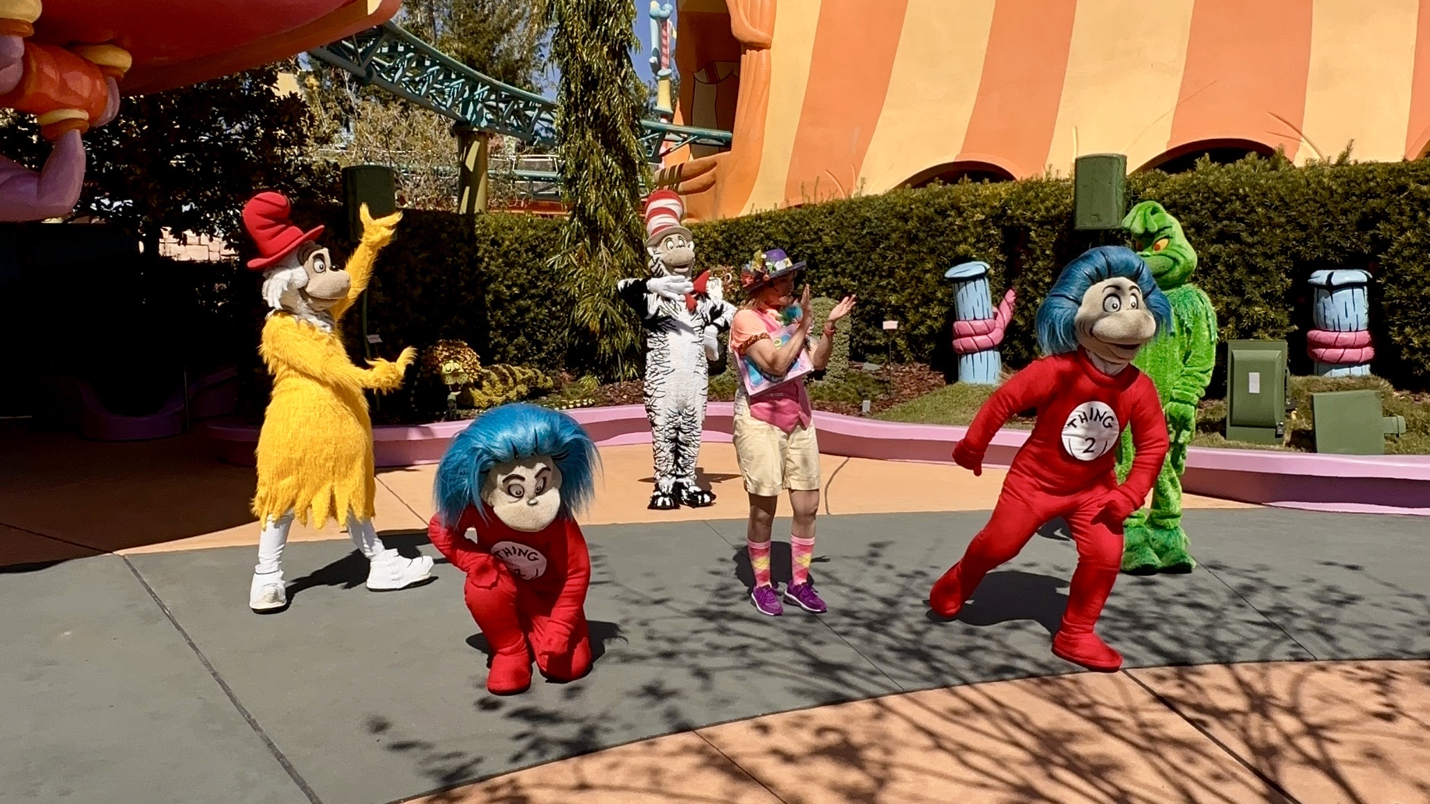 Characters in colorful costumes, including two in red outfits labeled "Thing 1" and "Thing 2," pose with a person in a hat at an outdoor theme park.
