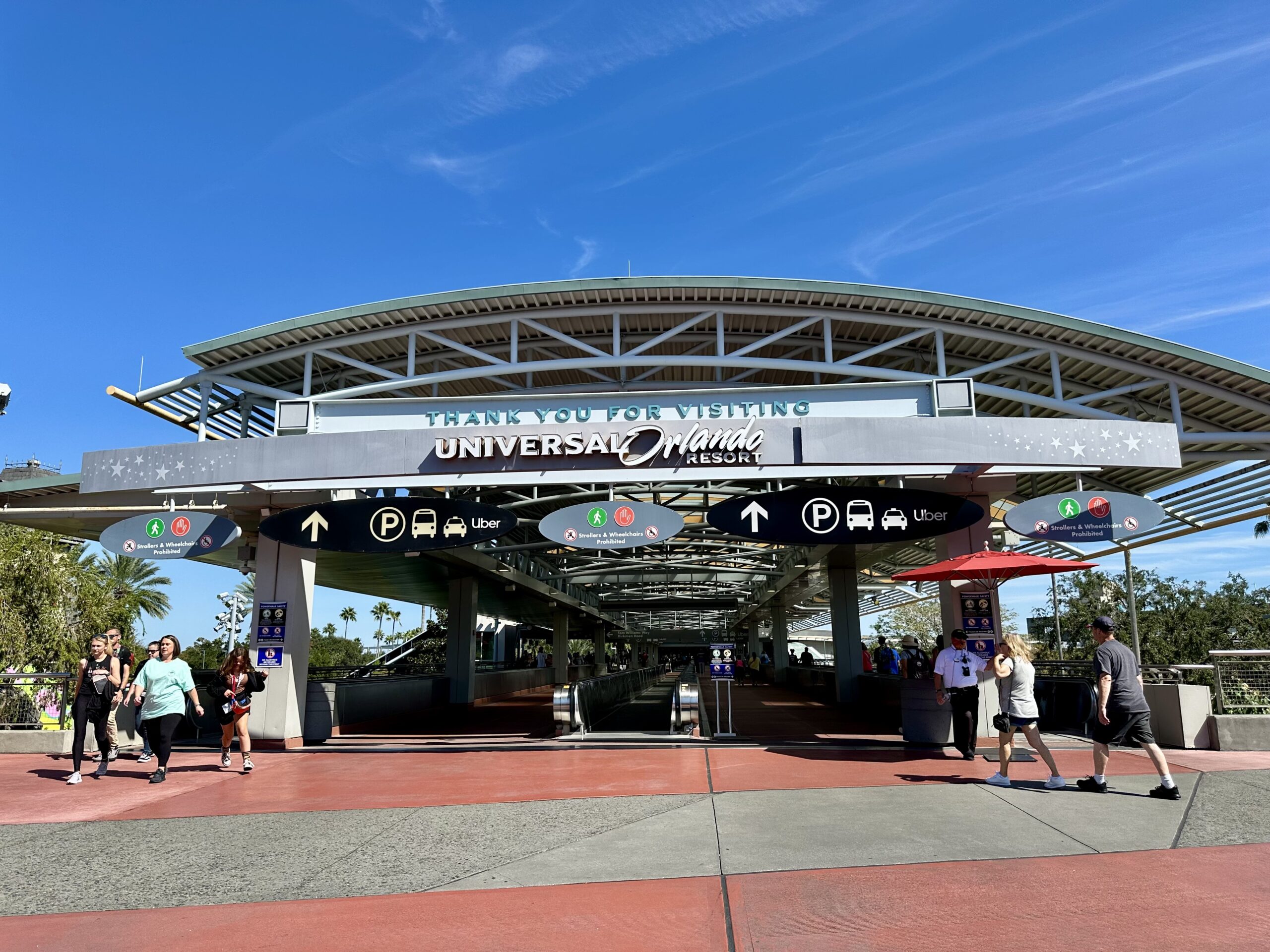 Entrance area of Universal Orlando Resort with people walking and signage for parking and transportation options. Clear blue sky above.