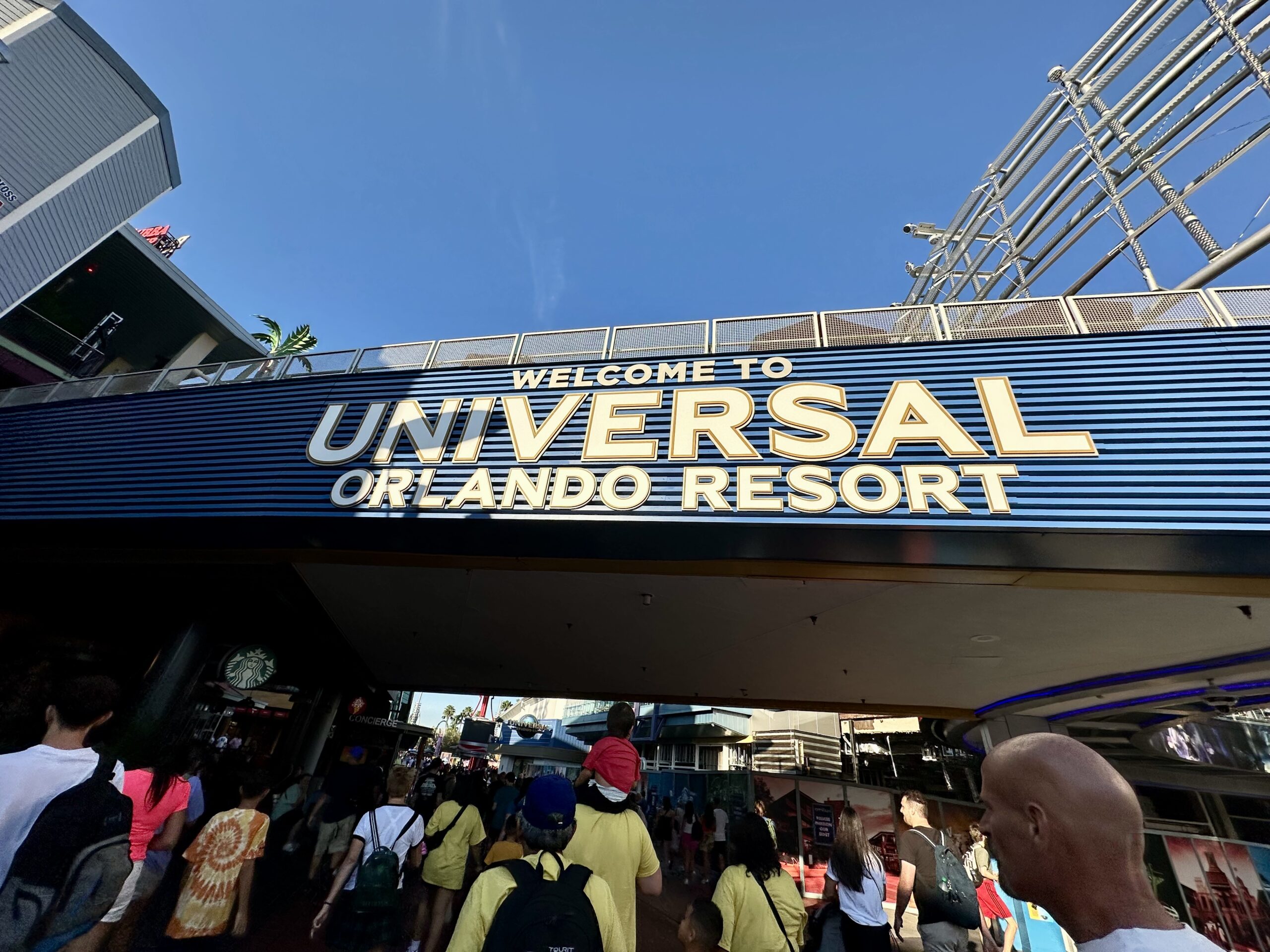 Entrance sign reading "Welcome to Universal Orlando Resort" with people walking below it.